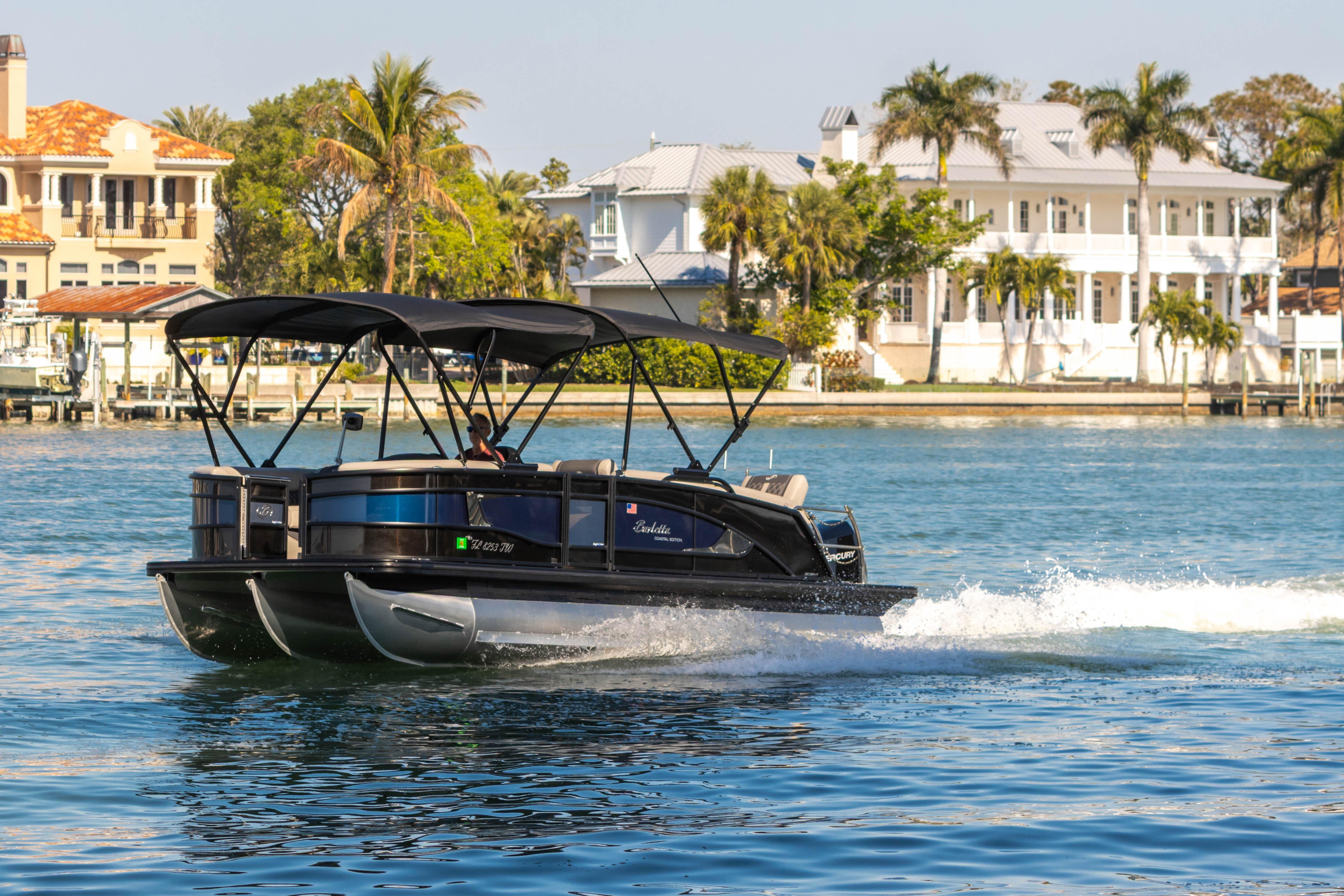 2024 Barletta L23U pontoon boat cruising on a scenic waterfront with luxury homes.