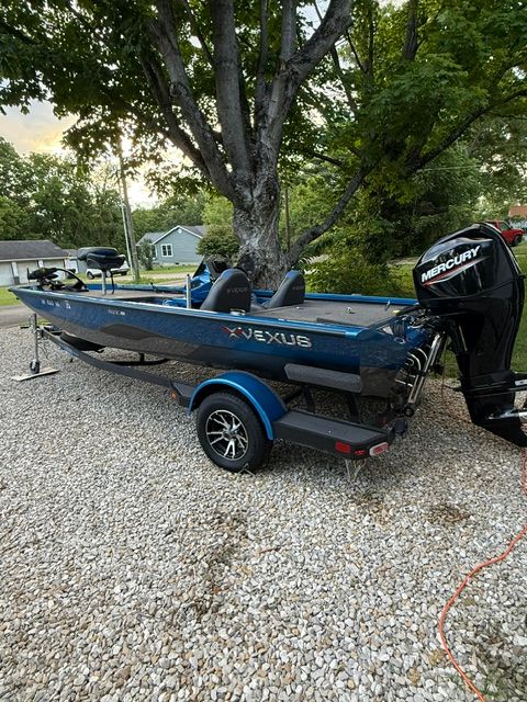 A blue Custom AVX181 boat on a trailer, parked on a gravel path next to trees.