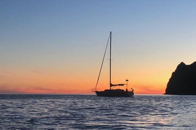 Sailboat Caliber 33 (1991) silhouetted against a vibrant sunset near rocky coastline.