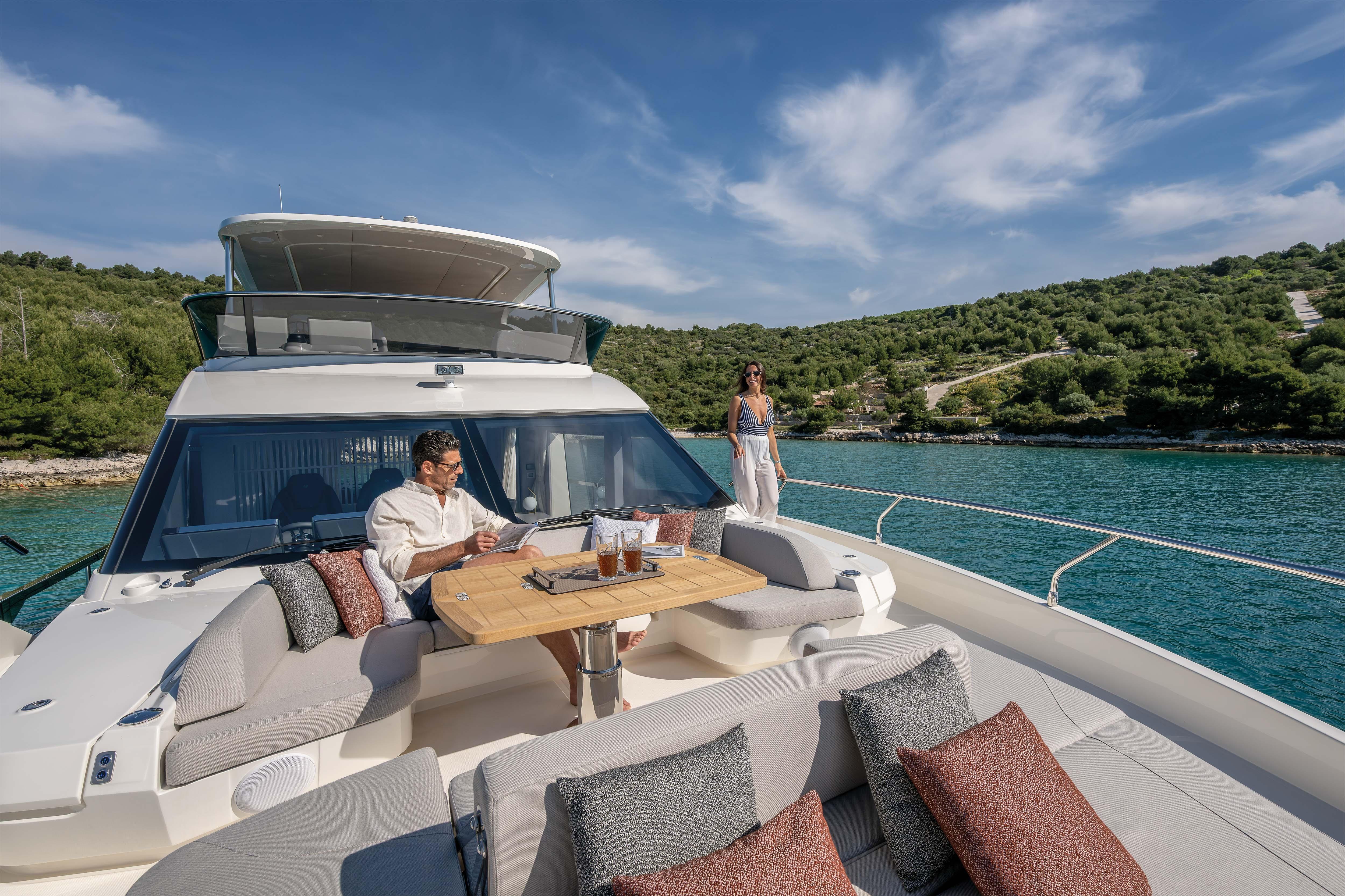 A luxurious boat with people relaxing on deck, surrounded by clear water and green hills.