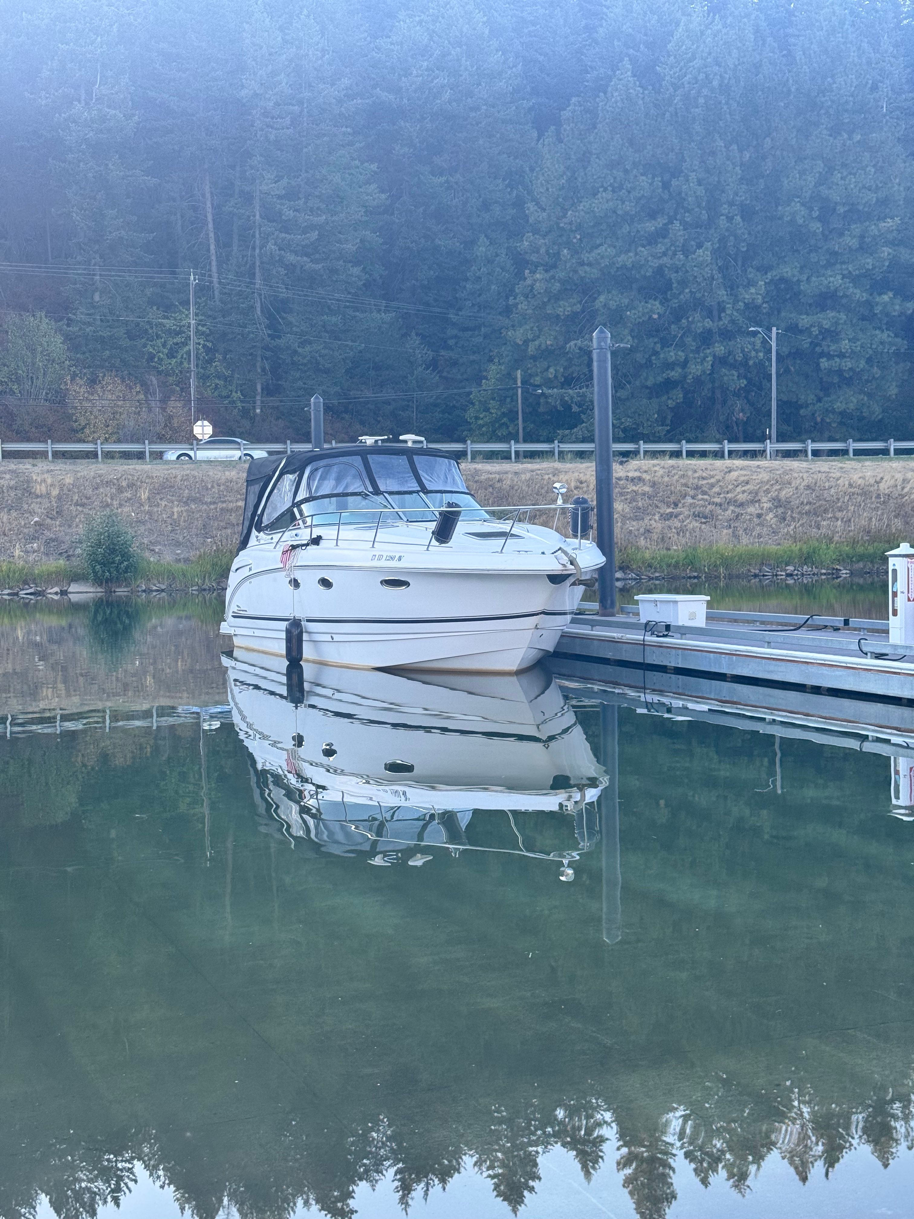 A white boat docked at a calm pier, surrounded by trees and still water.