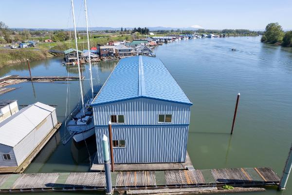 Blue boathouse on calm river, surrounded by docks and trees, under clear sky.
