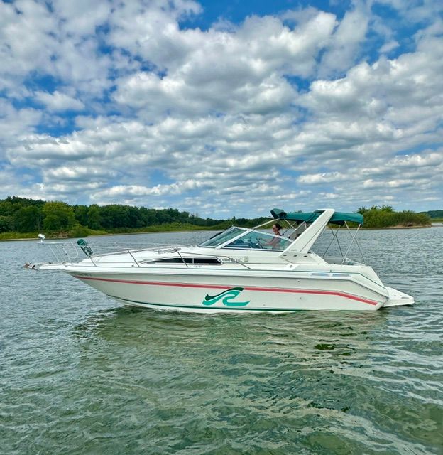 A white motorboat cruising on calm water, surrounded by trees and clouds.
