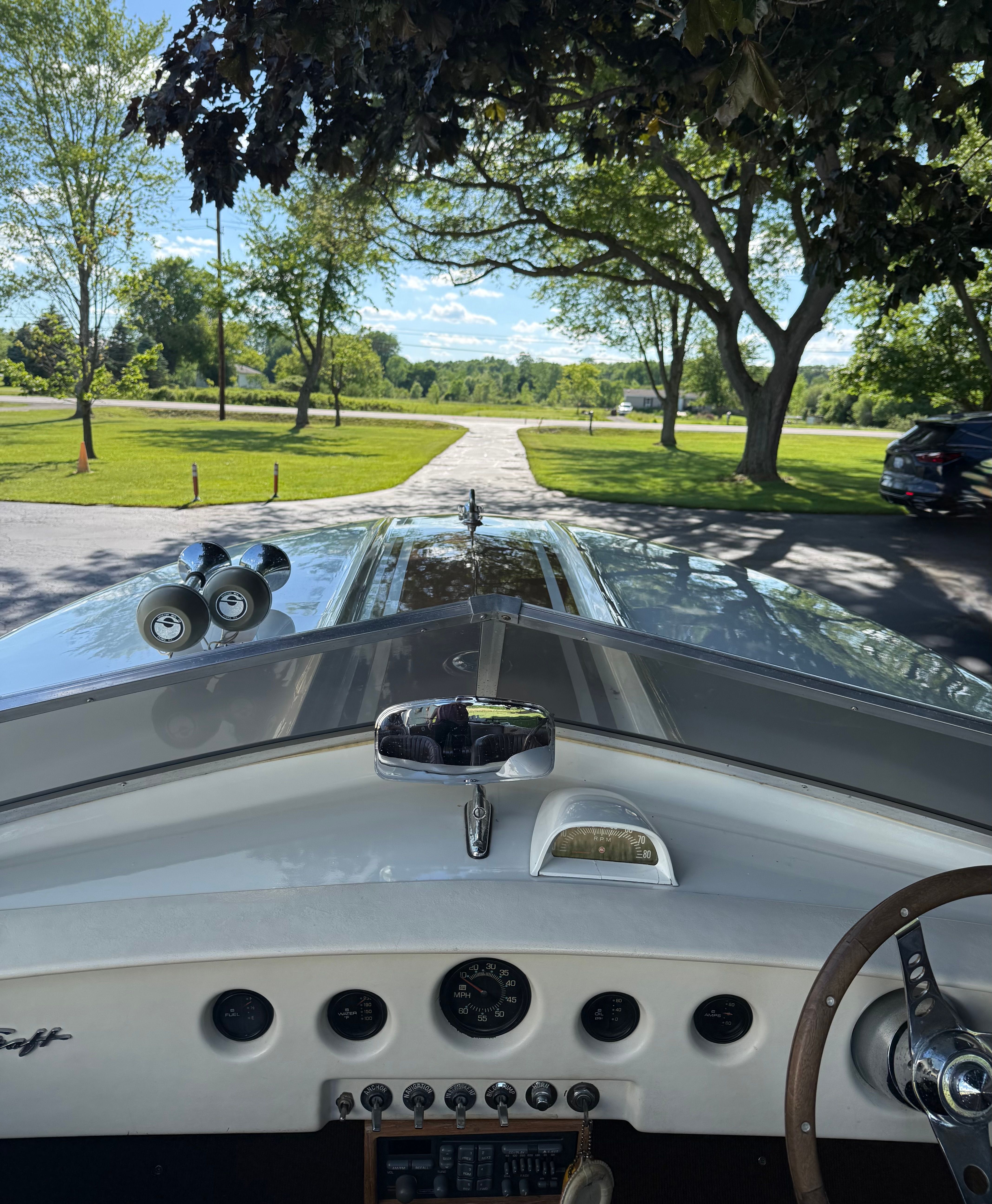 The image shows the cockpit of a 1972 Chris-Craft XK-19 boat, featuring a clear view of the dashboard and steering wheel. Trees and a sunny sky are visible in the background.