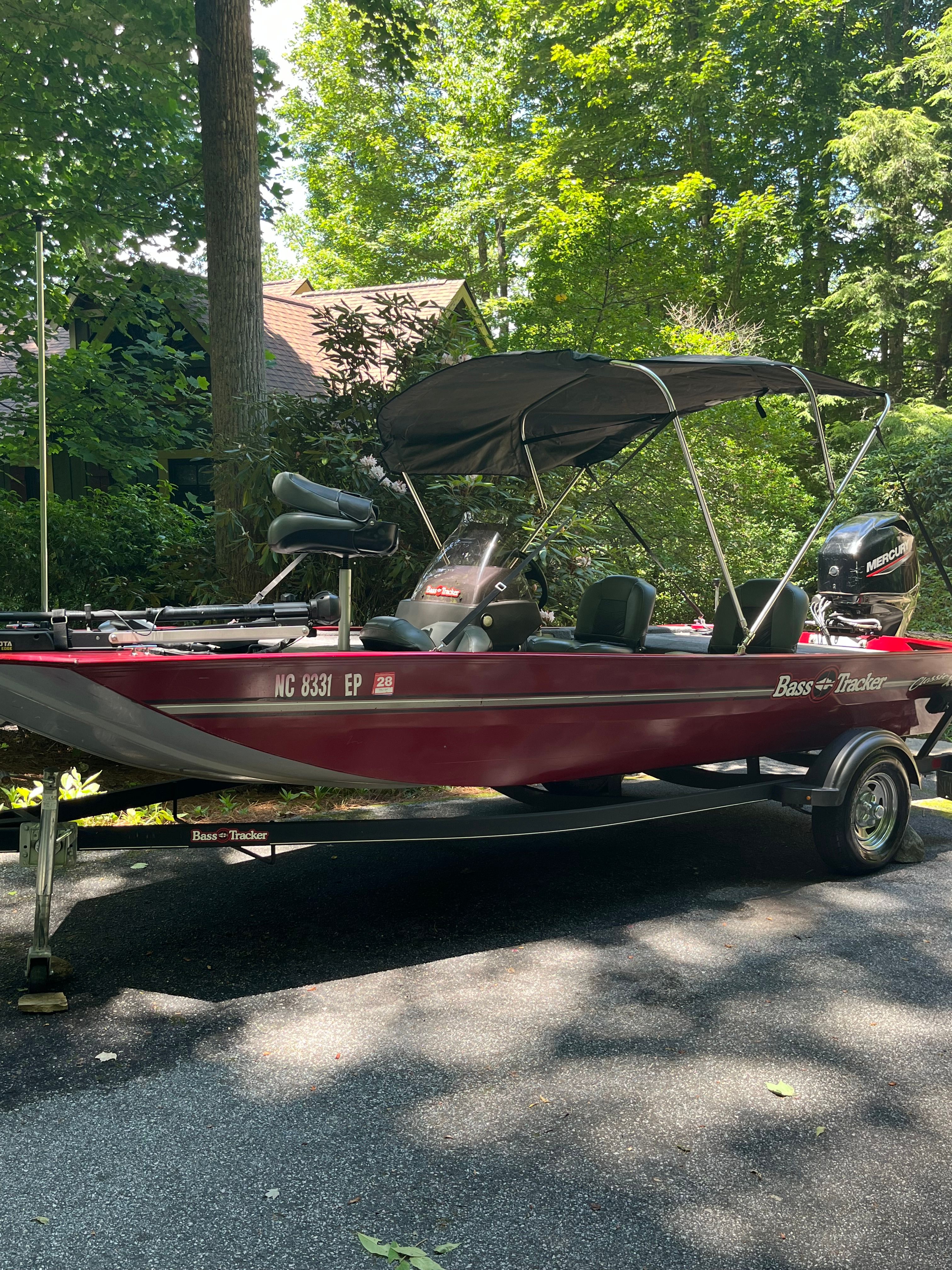 A red Tracker boat with a black canopy sits on a trailer in a wooded area.