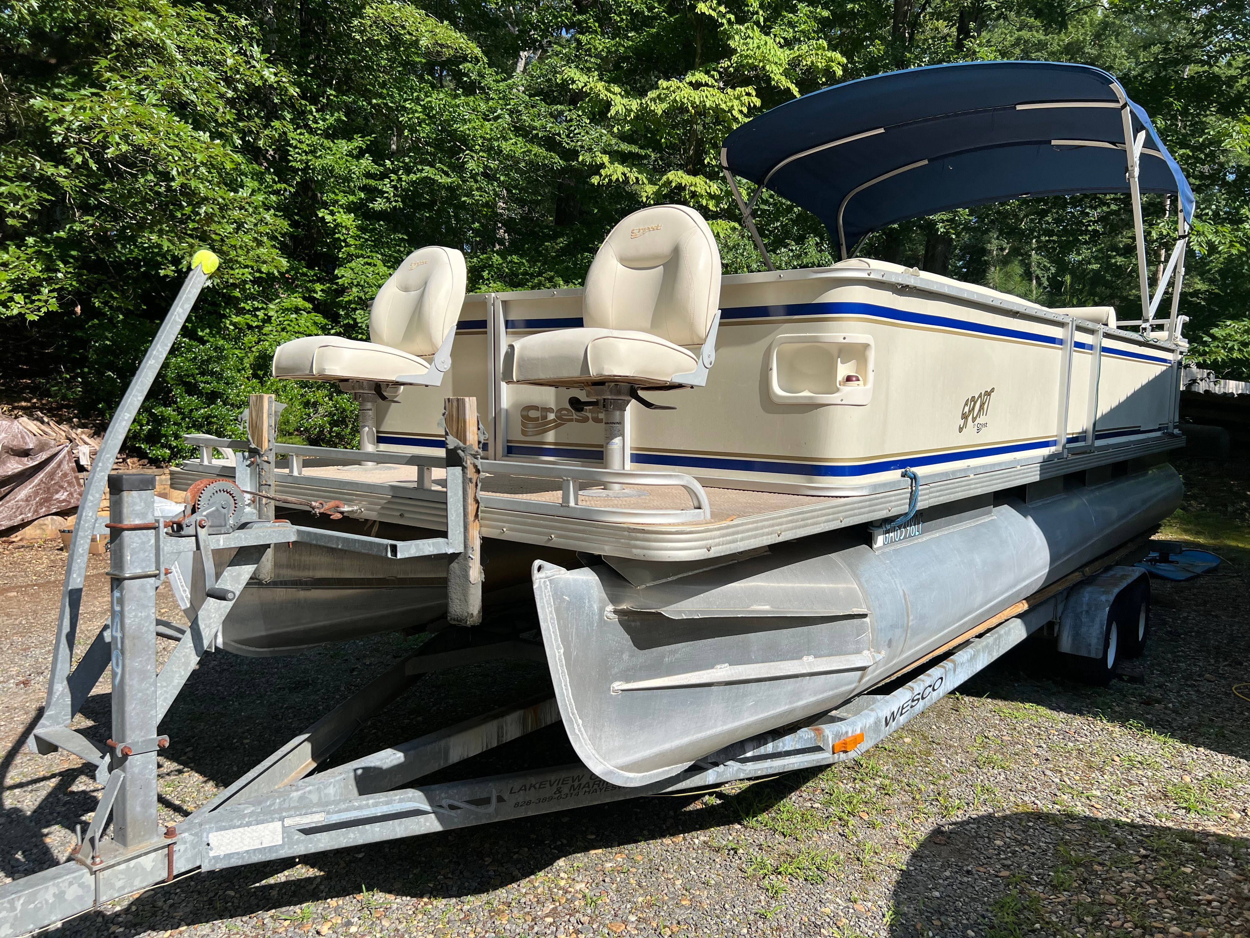A 2003 Crest 24’ Sport pontoon boat on a trailer, featuring two seats and a blue canopy.