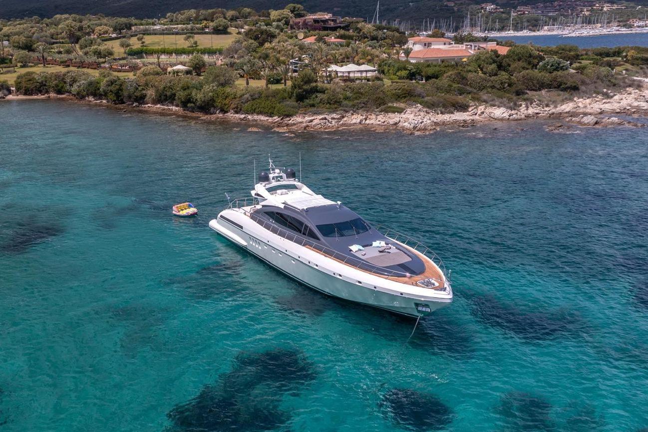 Luxurious 2006 Mangusta 92 yacht anchored in clear turquoise waters near a scenic coastline.