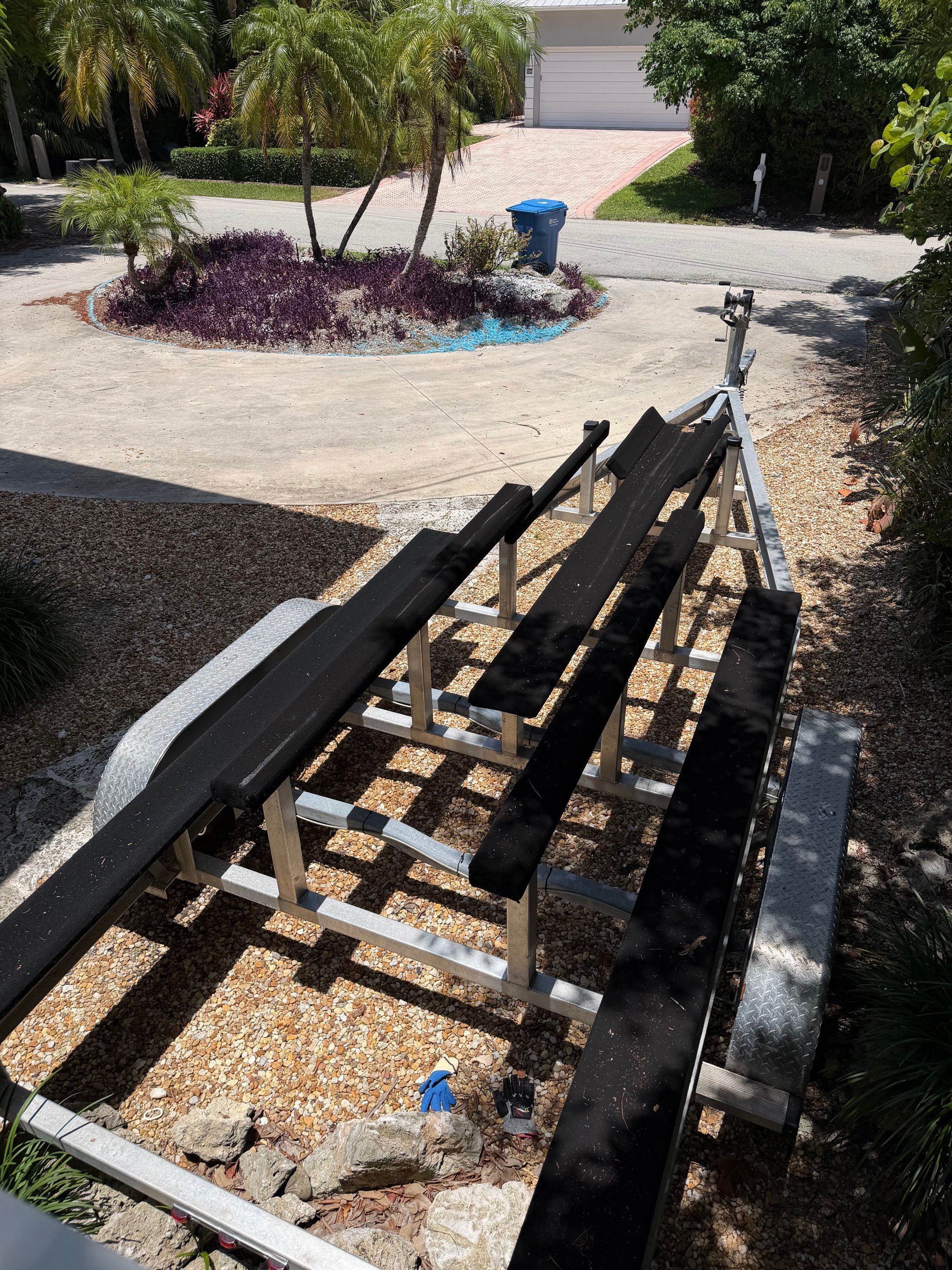 A boat trailer sits on a gravel driveway, surrounded by palm trees and shrubs.