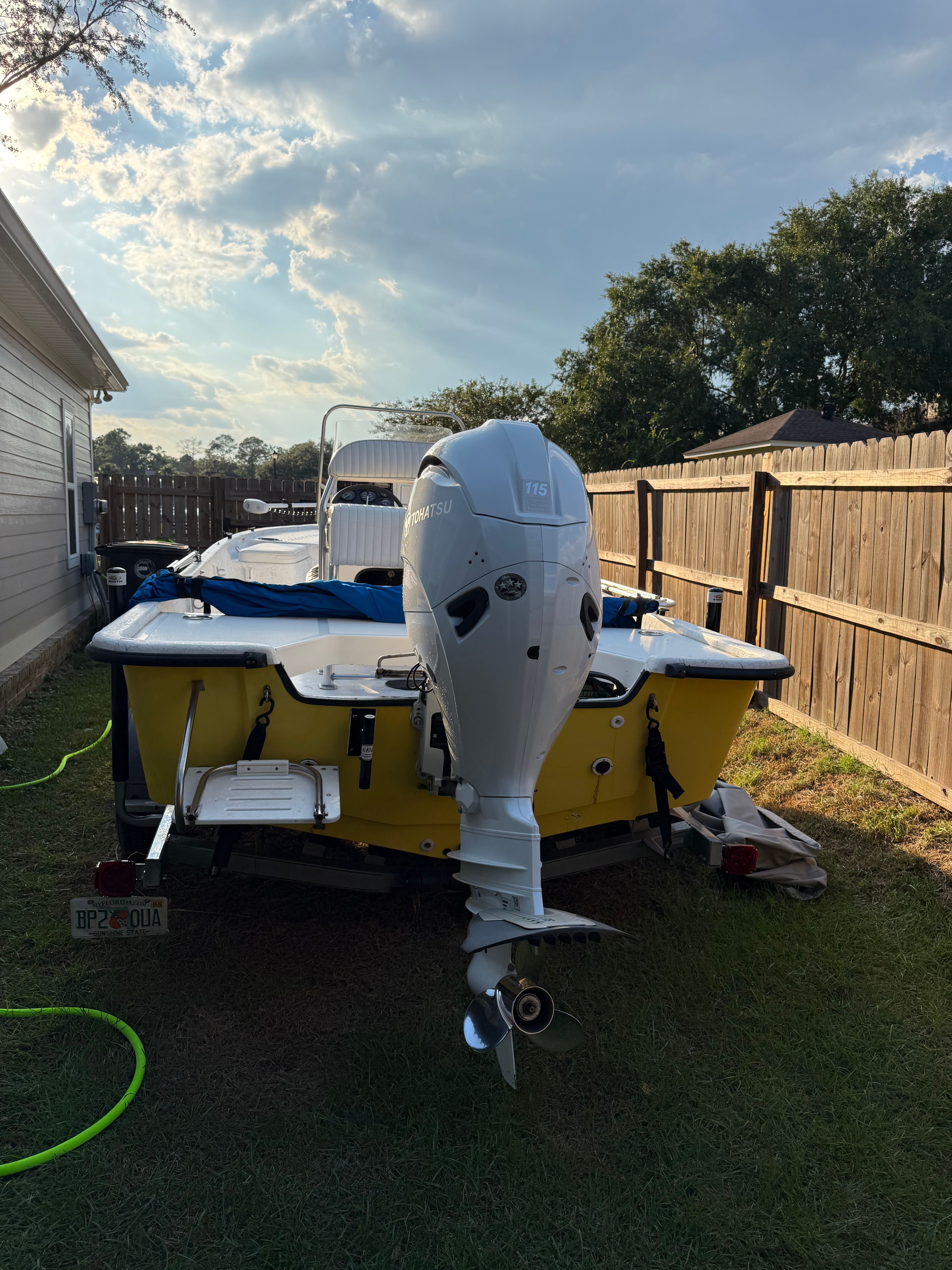 A yellow boat with an outboard motor is on a trailer in a backyard.