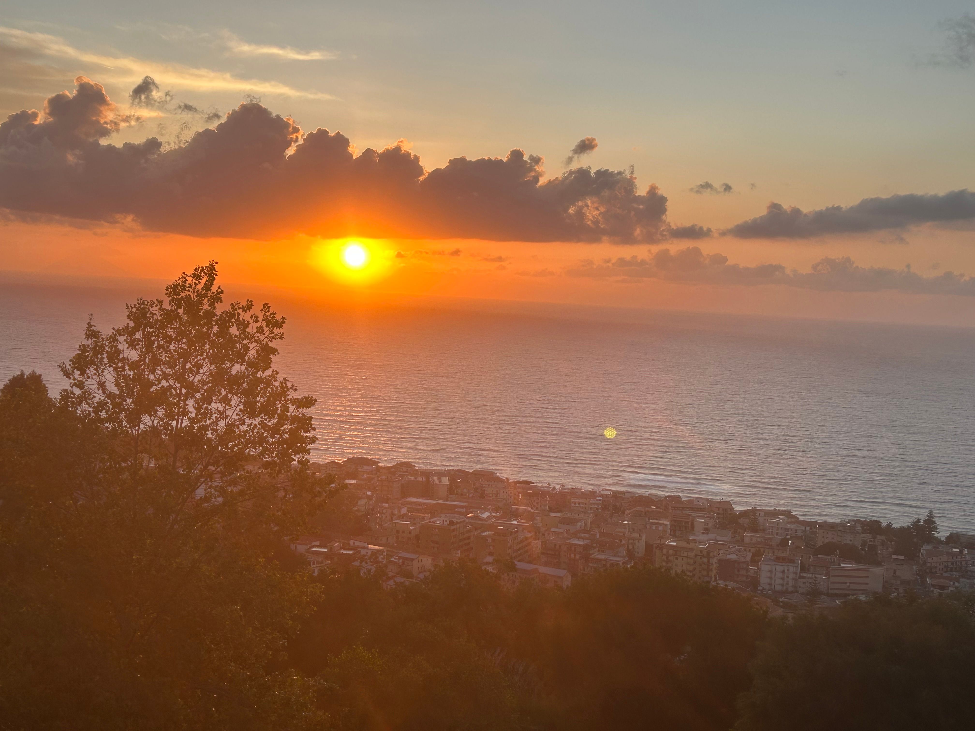 Sunset over the sea with a view of a coastal town and trees in the foreground.