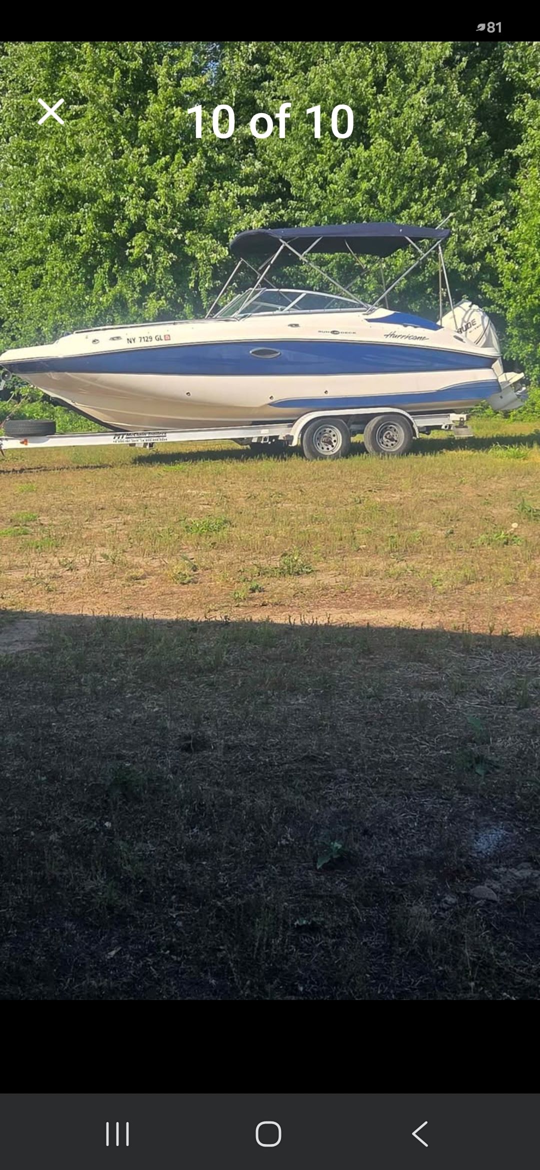 A white boat with a blue canopy sits on a trailer in front of green trees.