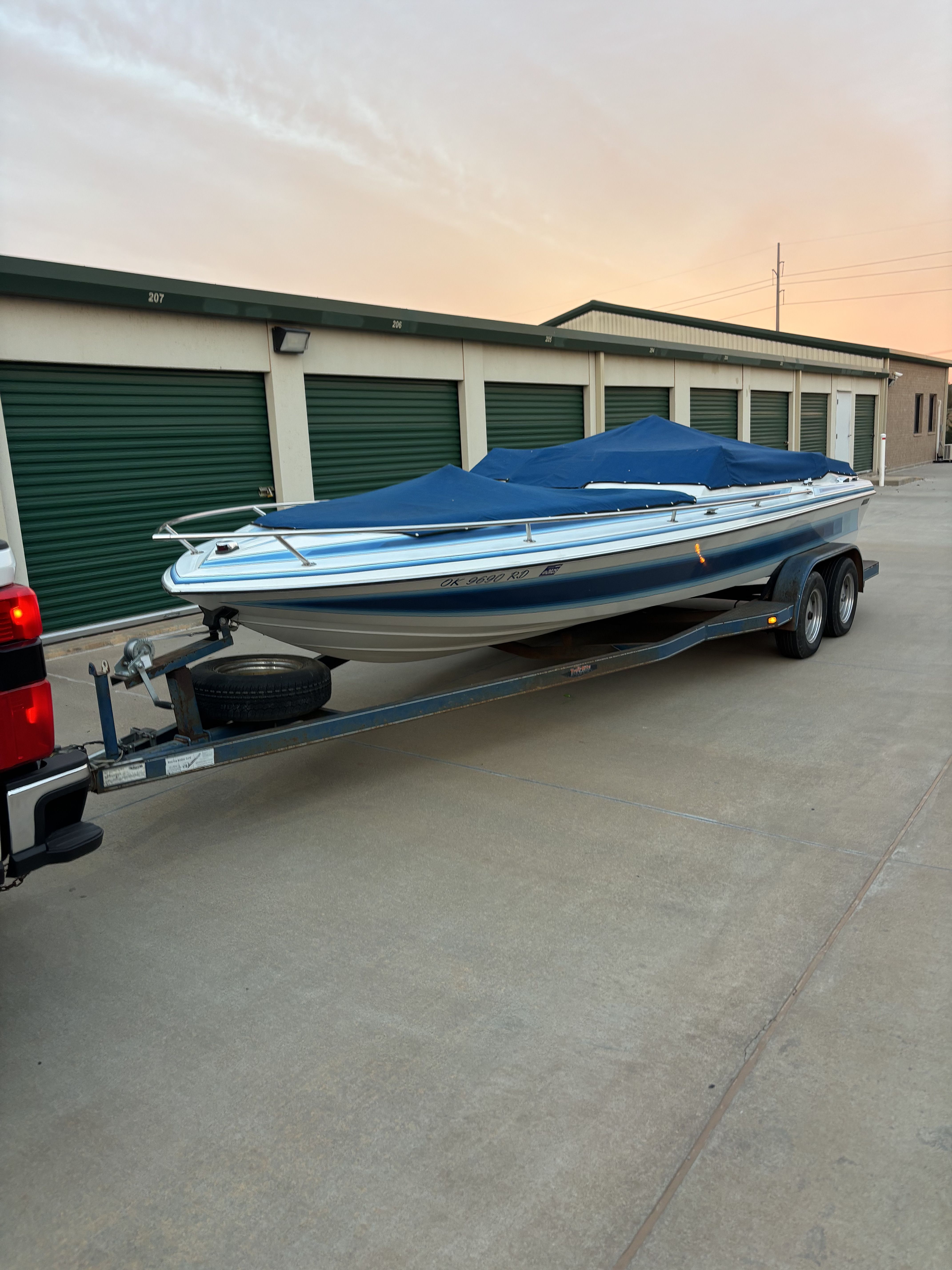 A blue boat covered with a tarp is on a trailer in front of storage units.