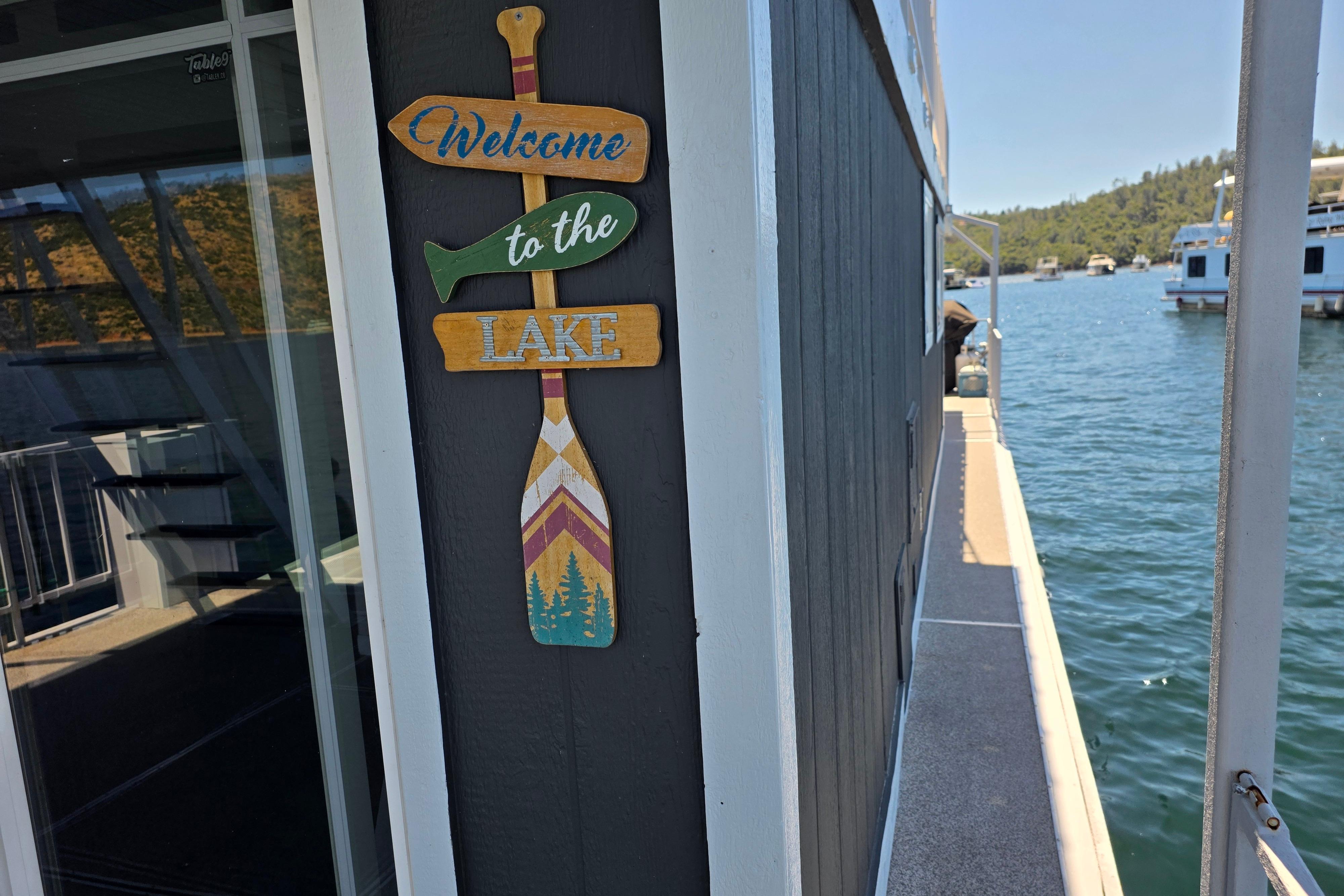 Houseboat with "Welcome to the Lake" sign, docked on a scenic lake, 2007 Custom model.