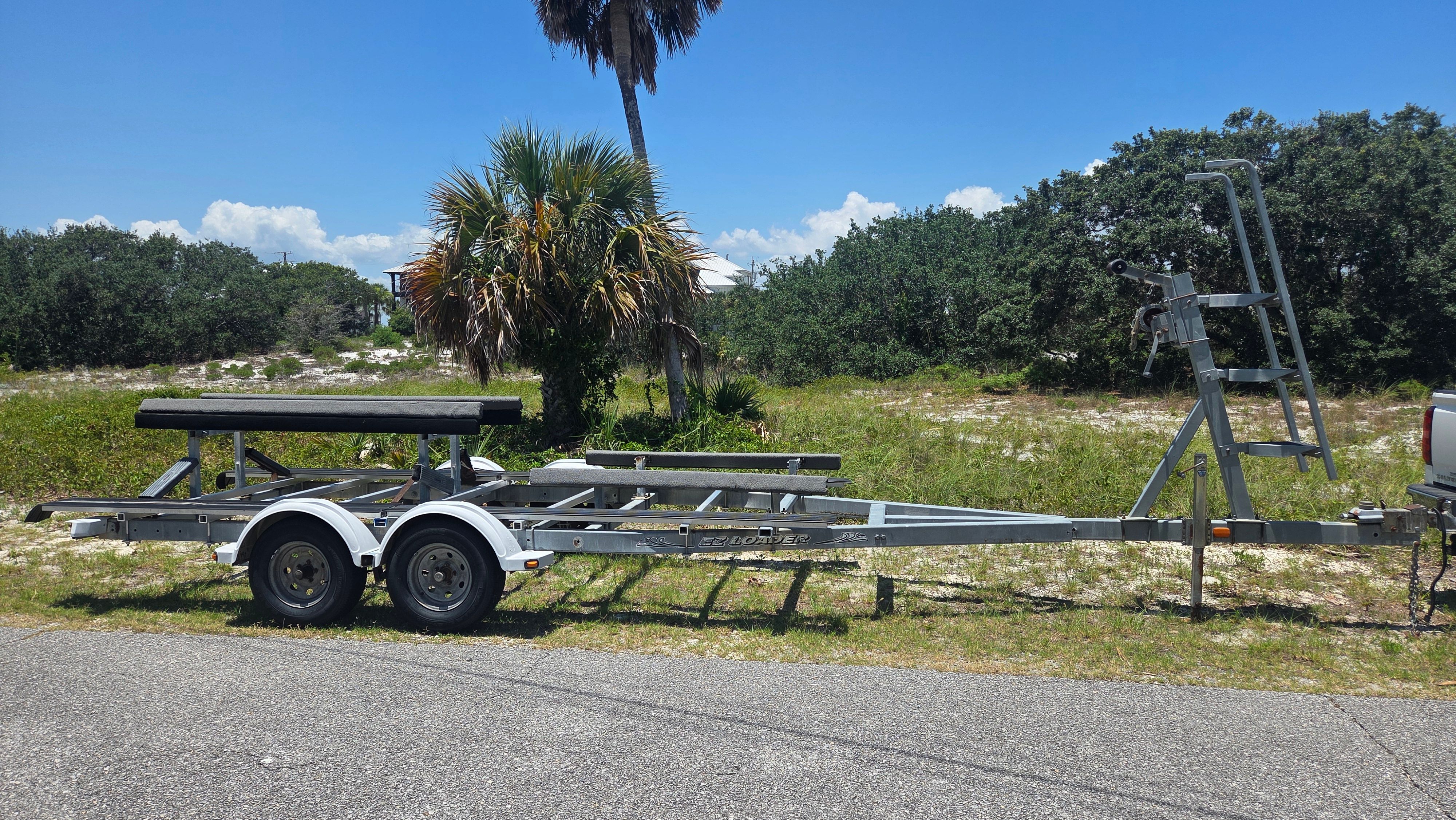 A boat trailer sits on a road, surrounded by palm trees and bushes.