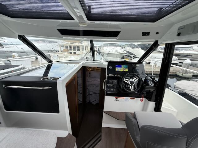 Interior view of a modern boat helm with gray seating and a large steering wheel. Windows overlook the marina.