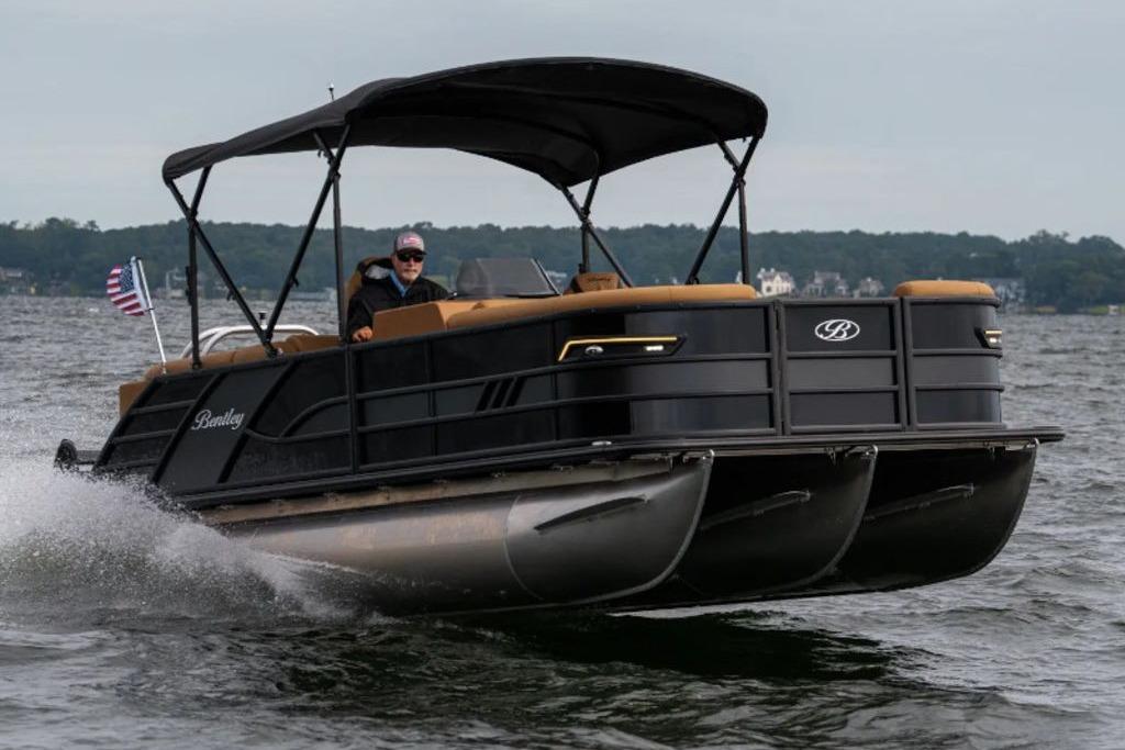 2025 Bentley Pontoons Elite 253 Admiral Tri-toon cruising on a lake with American flag.