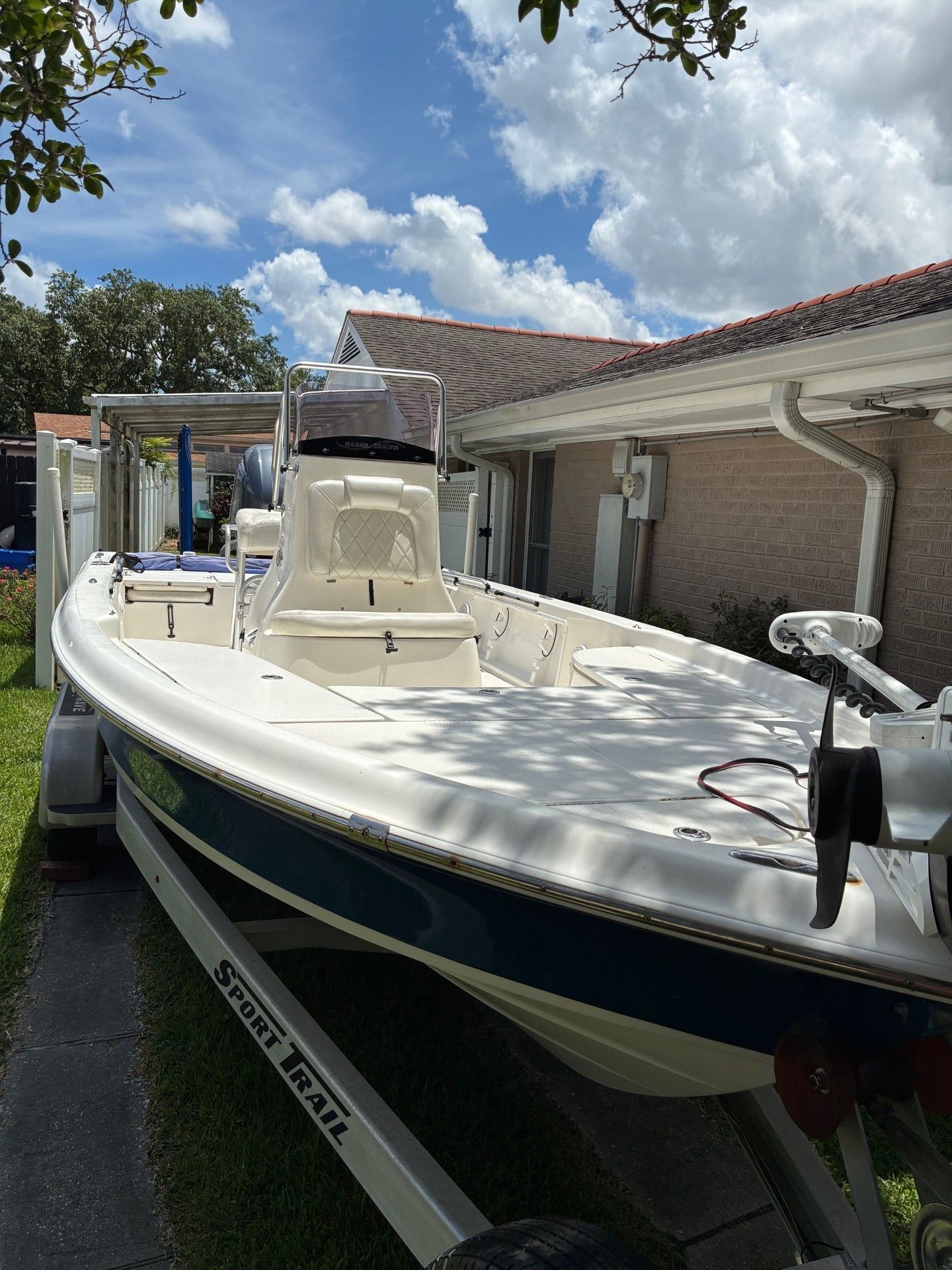 A white boat on a trailer in a yard, surrounded by trees and a house.