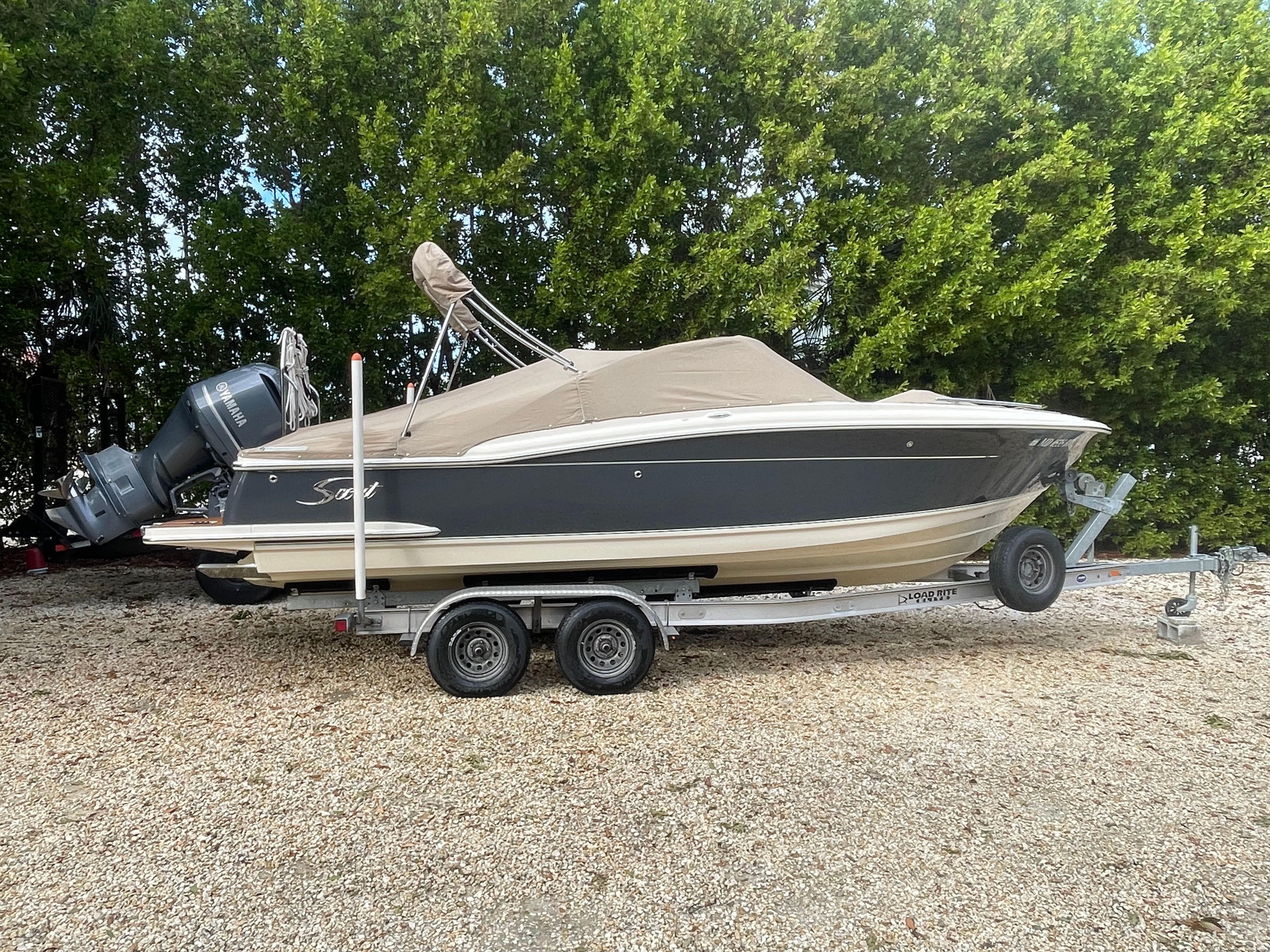 Scout 235 Dorado 2020 on a trailer, covered, parked on gravel in front of trees.
