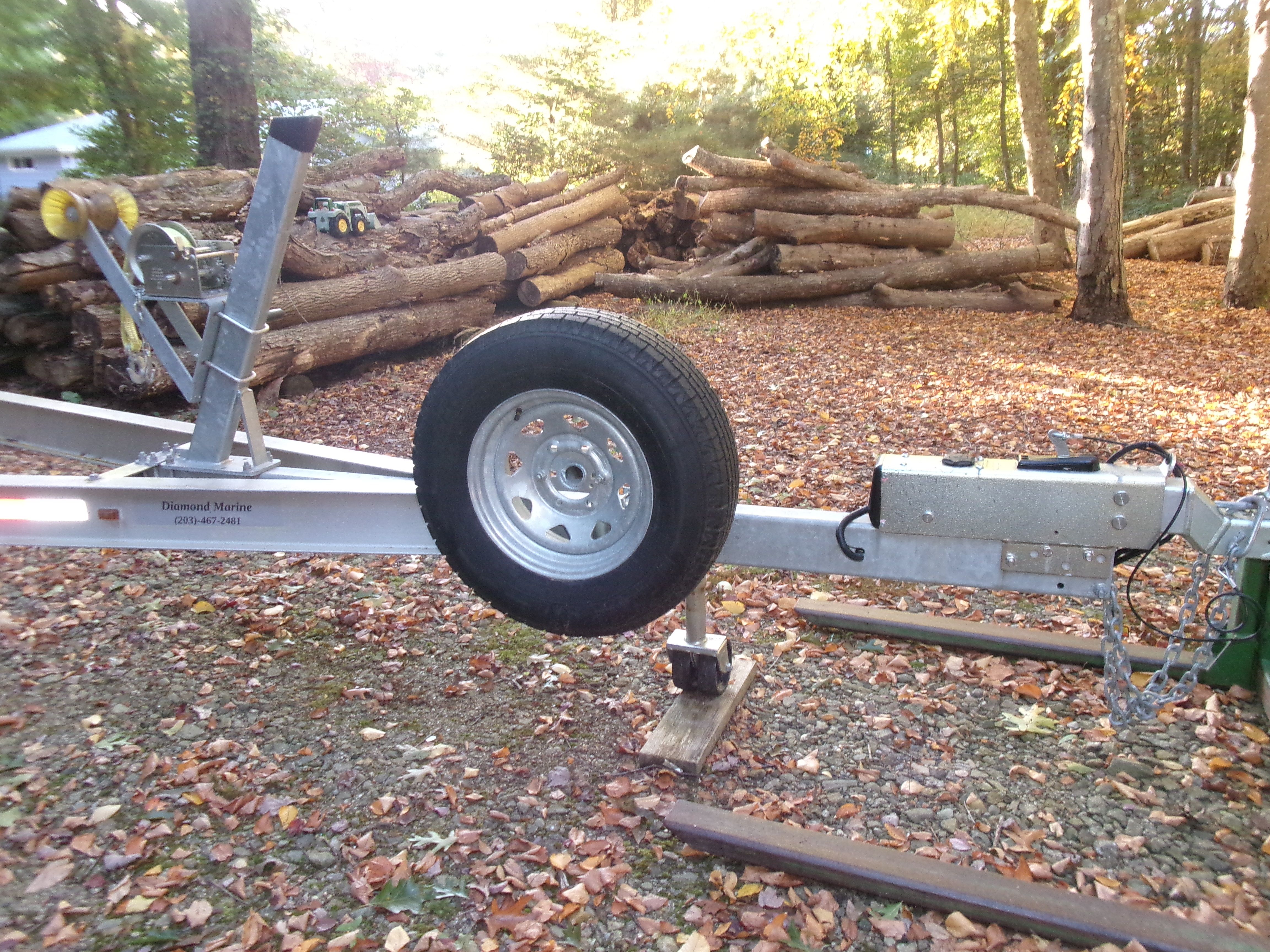 A boat trailer sits on a leaf-covered ground, surrounded by stacked logs.