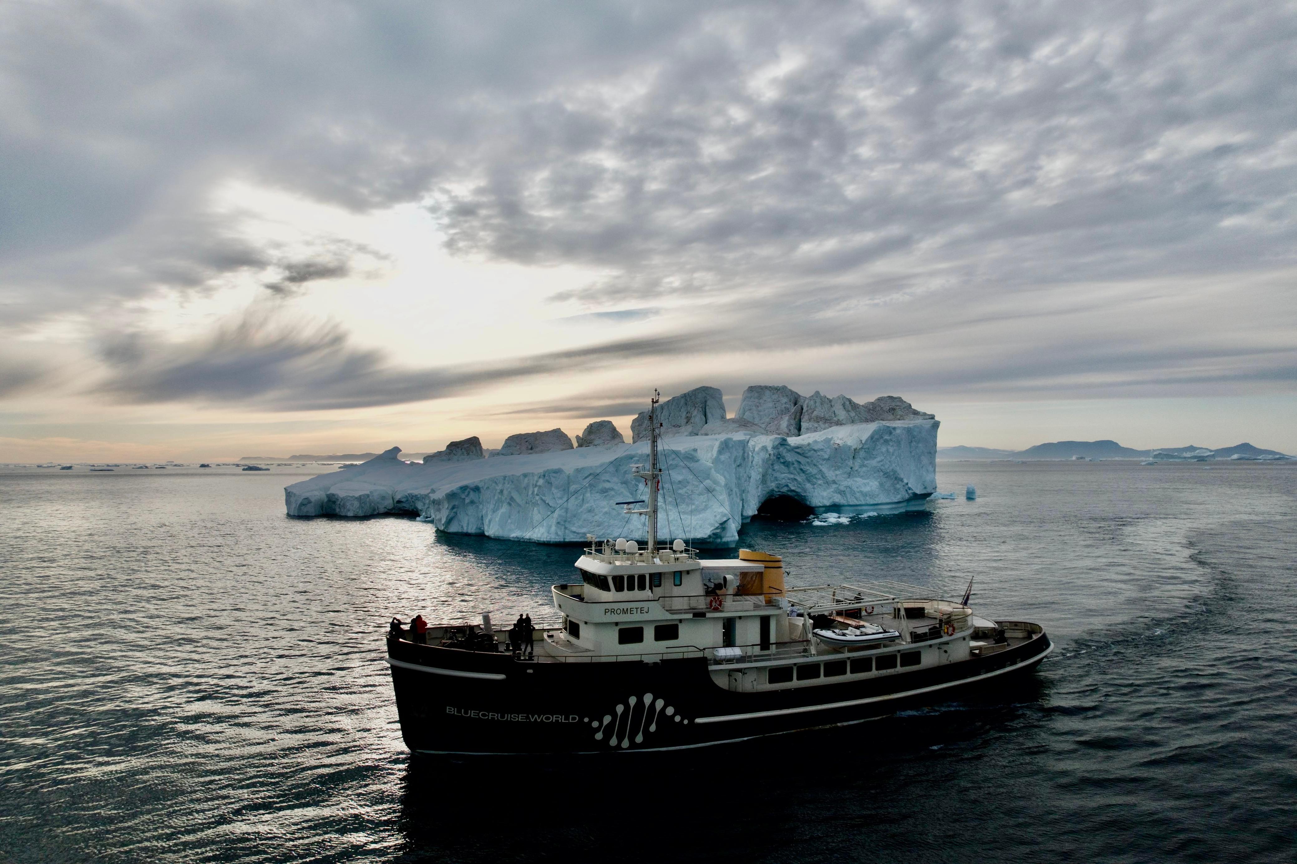 A 1956 custom converted tugboat navigates near a large iceberg under a cloudy sky.