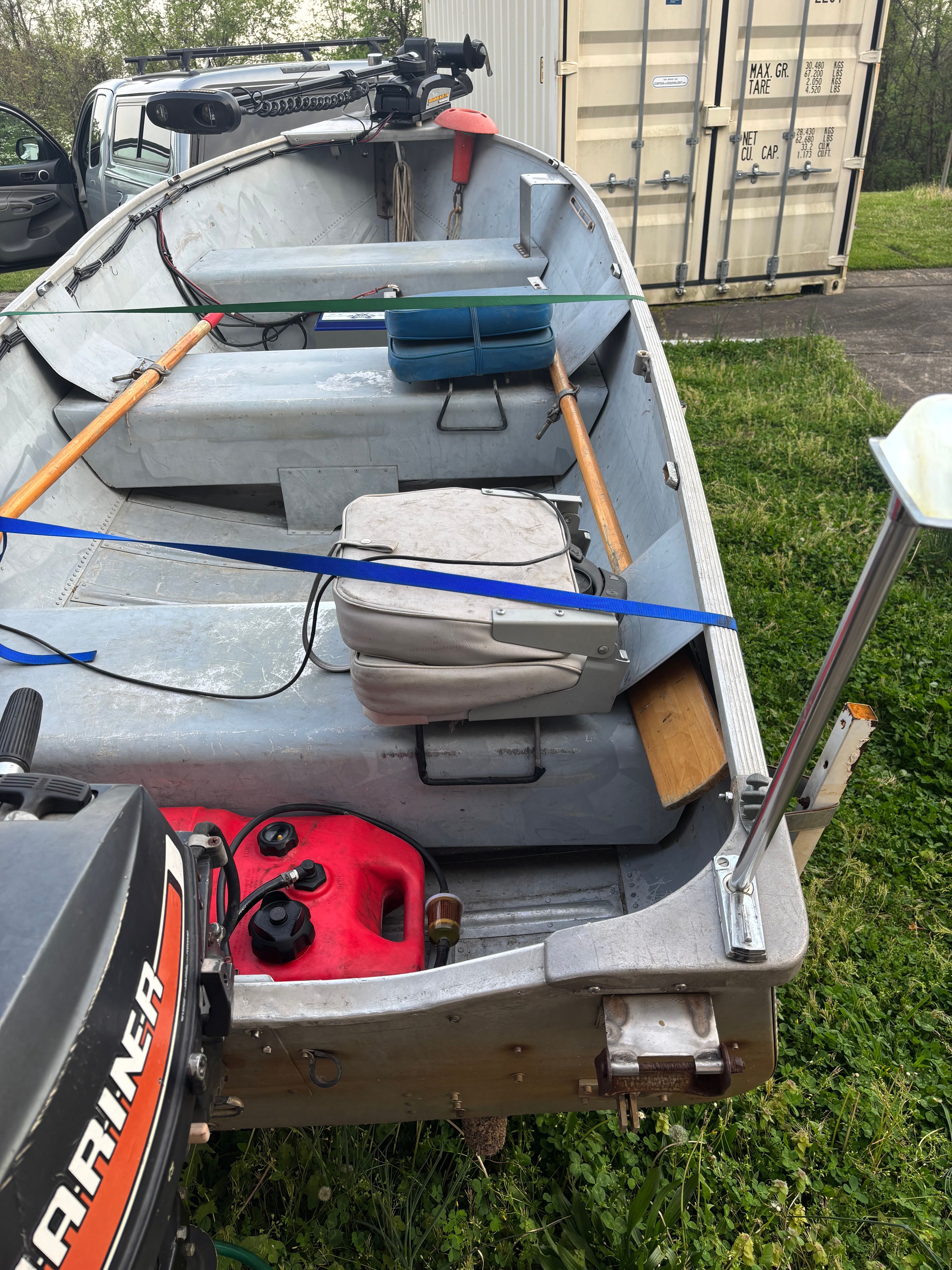 A small aluminum boat with oars and motor, on grass next to a container.