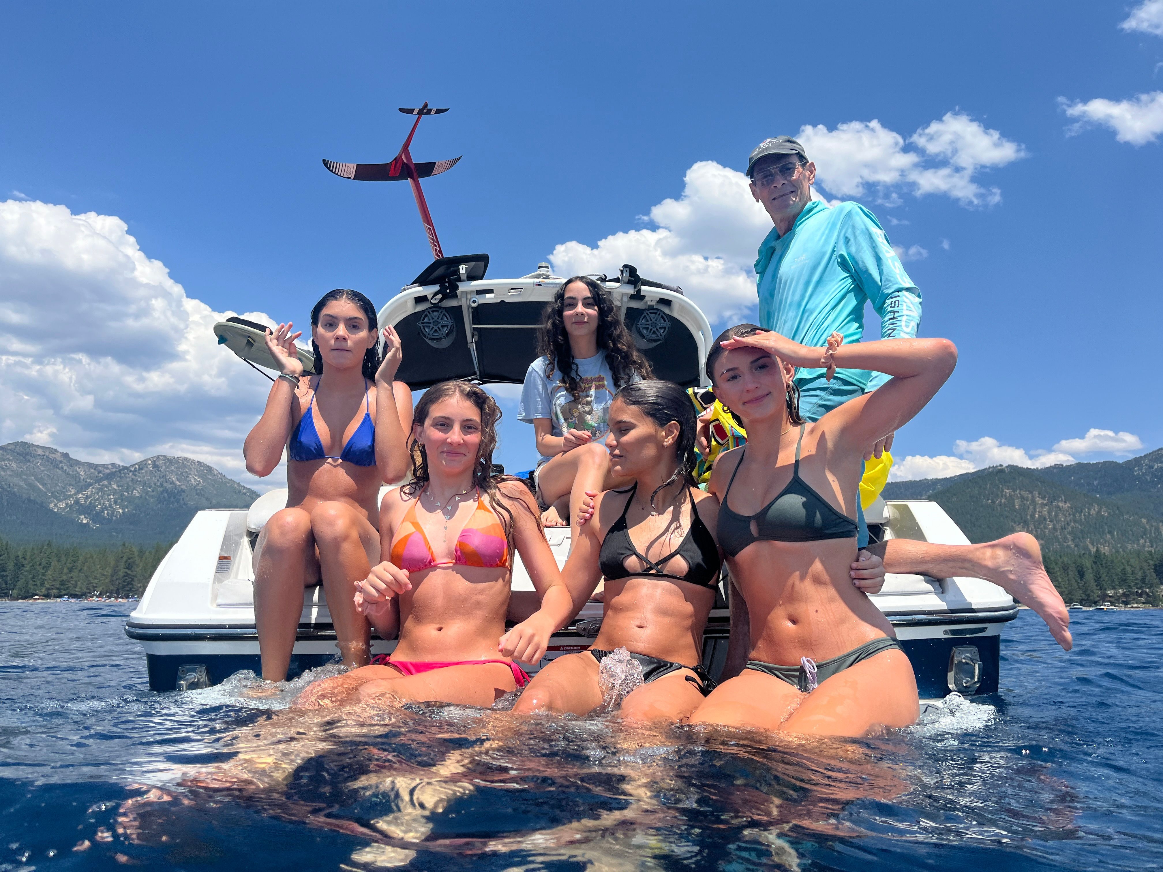 A group of people sits on a boat in the water, surrounded by mountains and blue sky.
