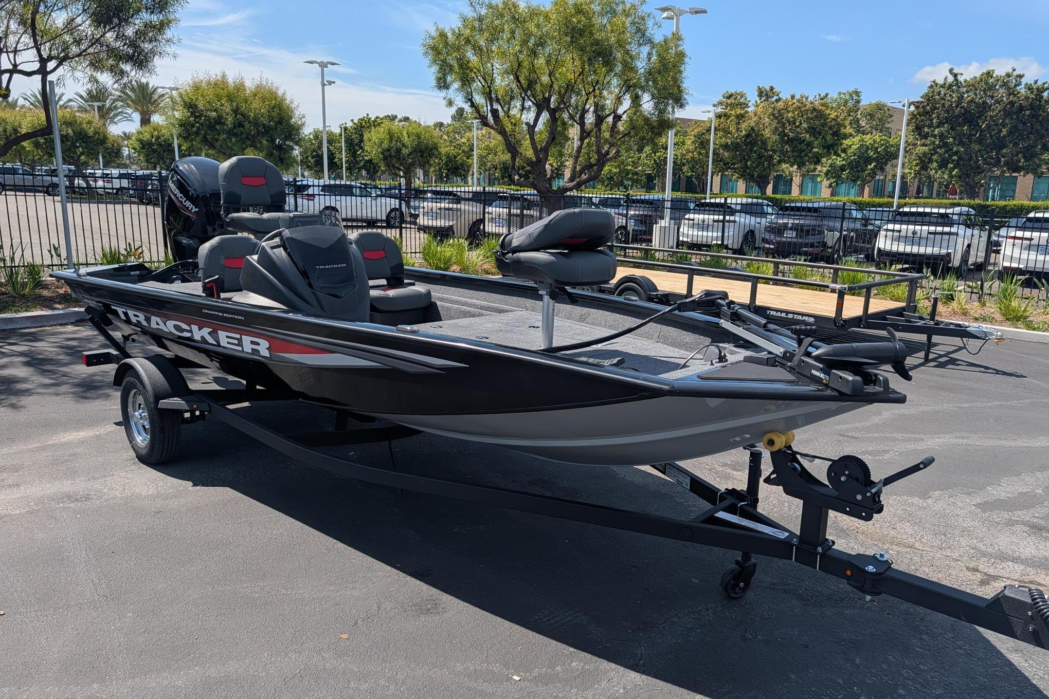 A black Tracker boat on a trailer, surrounded by trees and cars in a parking lot.