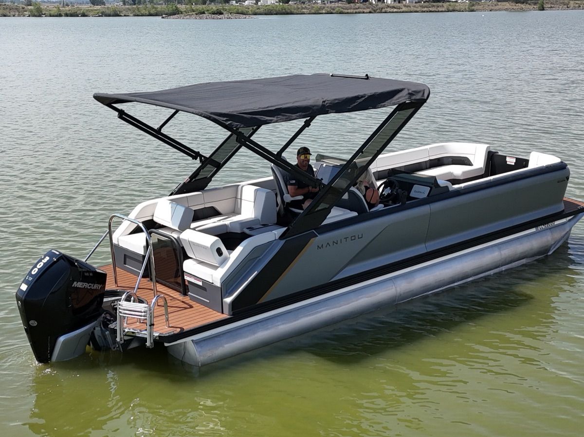 A 2024 Manitou Explore pontoon boat on a calm lake with a black canopy.