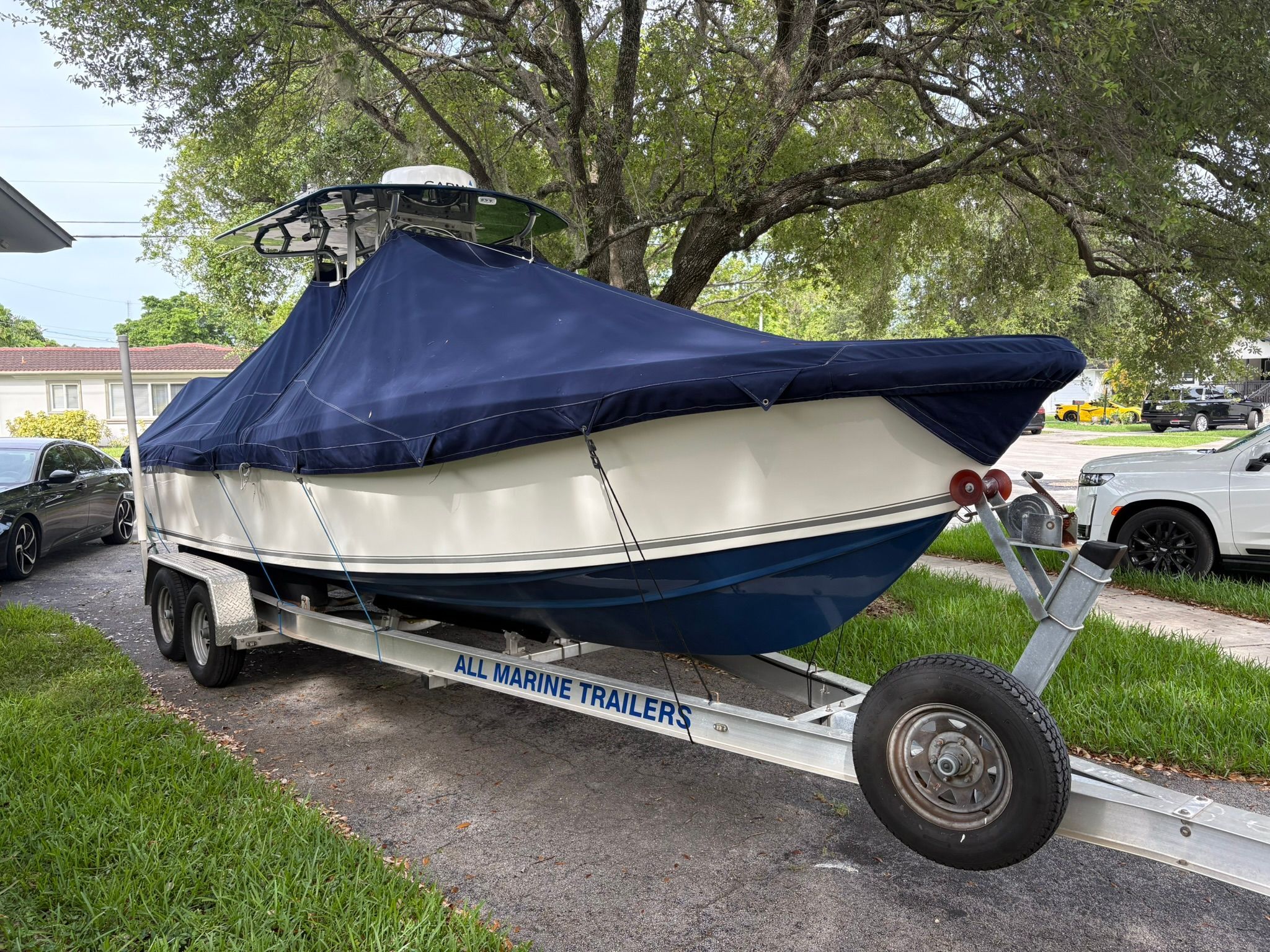 A white boat with a blue cover is on a trailer in a residential area.