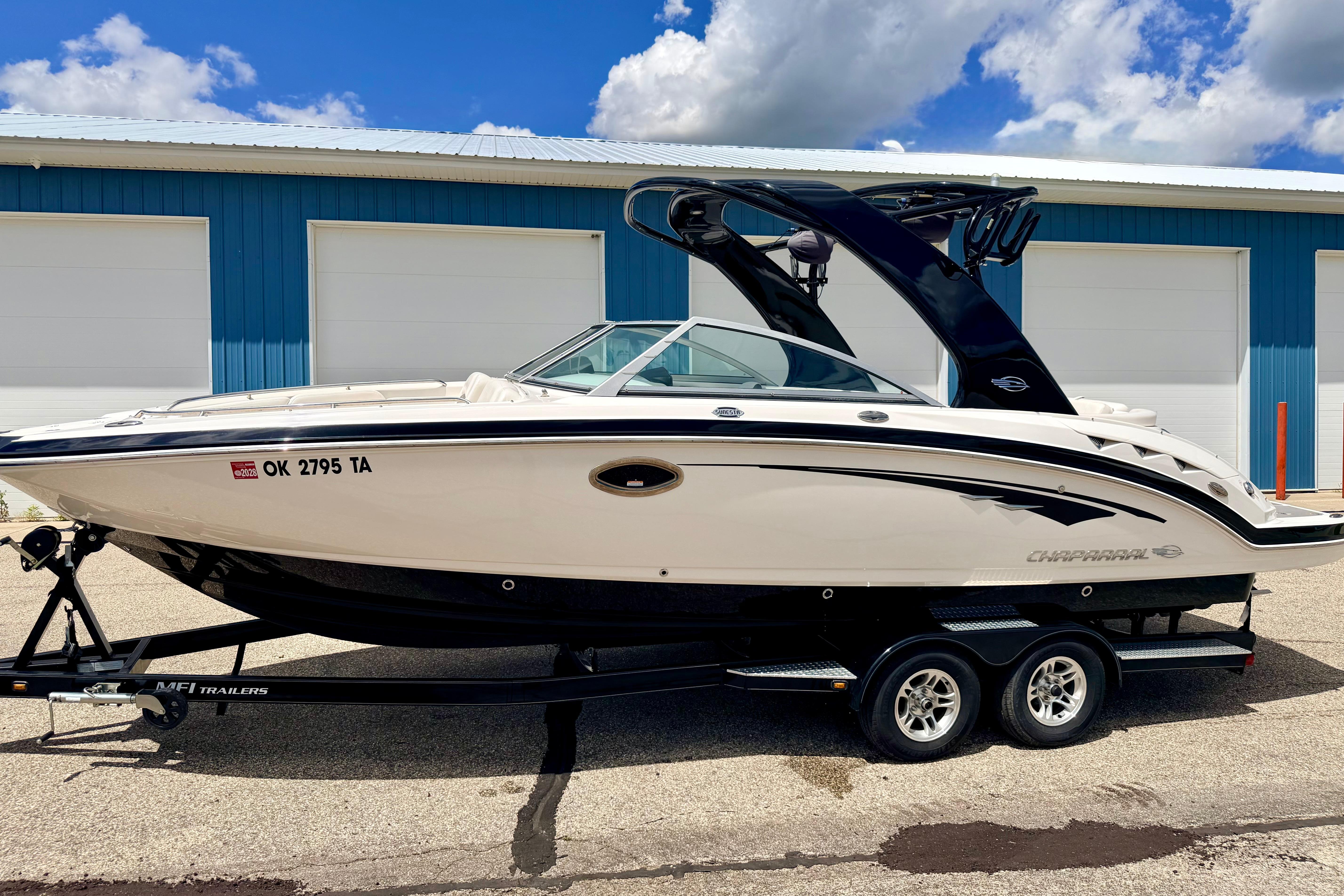 2015 Chaparral Sunesta 264 boat on trailer, parked outside blue garage under cloudy sky.