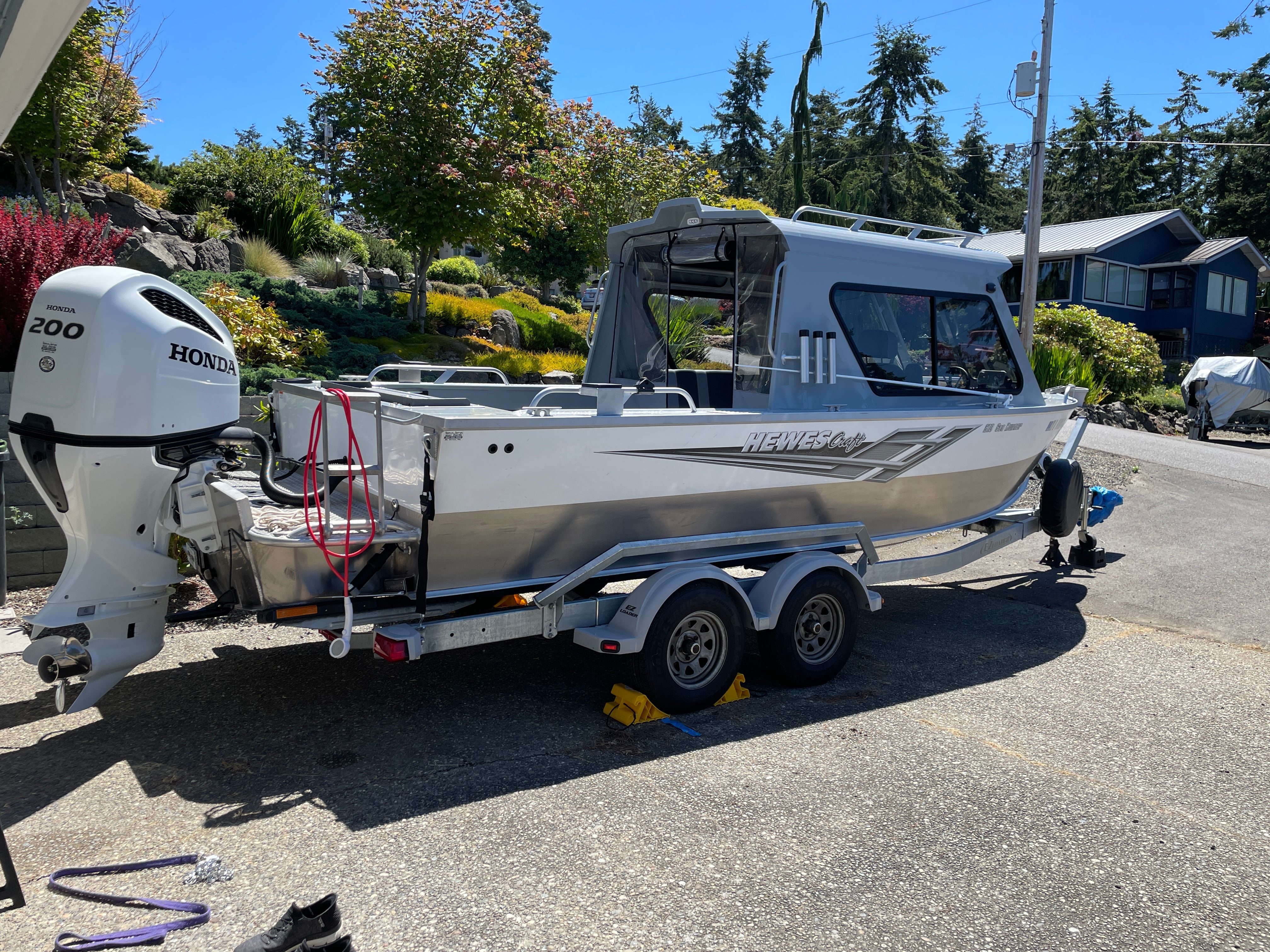 A silver hardtop boat on a trailer, surrounded by trees and houses.