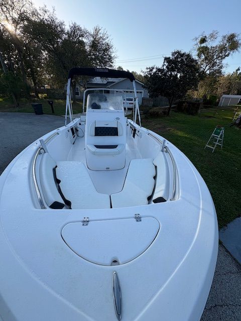A white boat with an open deck and seating, surrounded by trees and a house in the background.