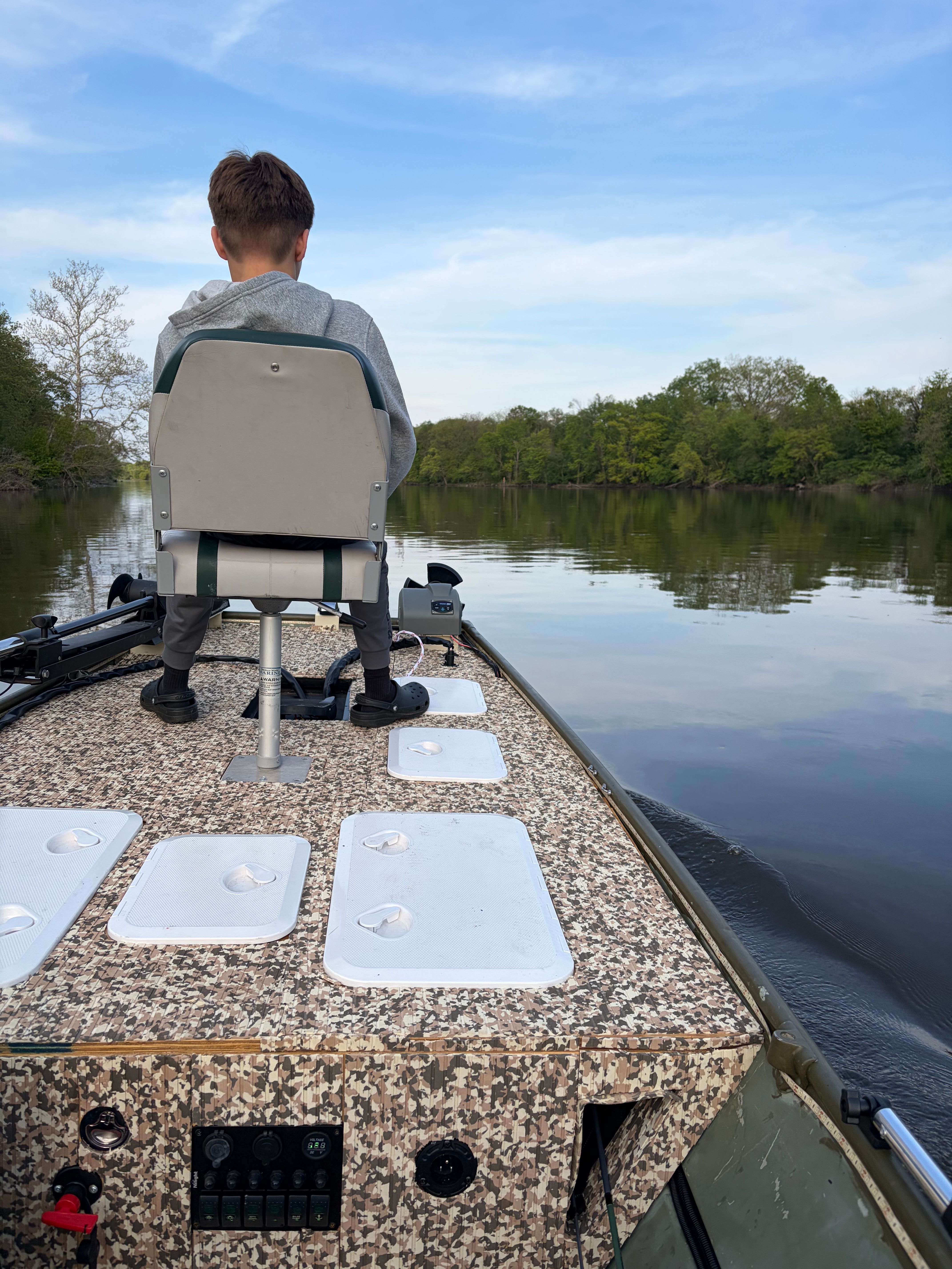 A boy sits on a chair on a boat cruising a calm river.