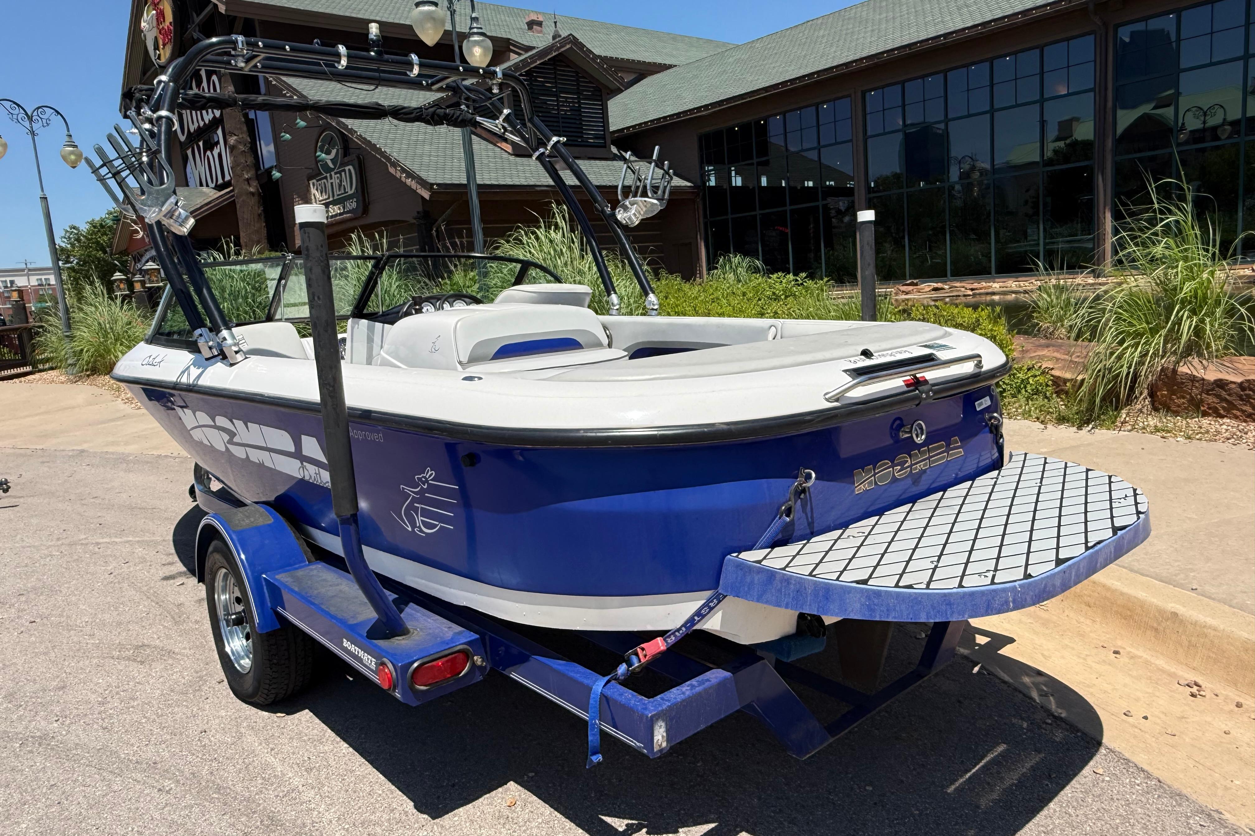 2001 Moomba Outback boat with blue hull, parked on a trailer near a building.