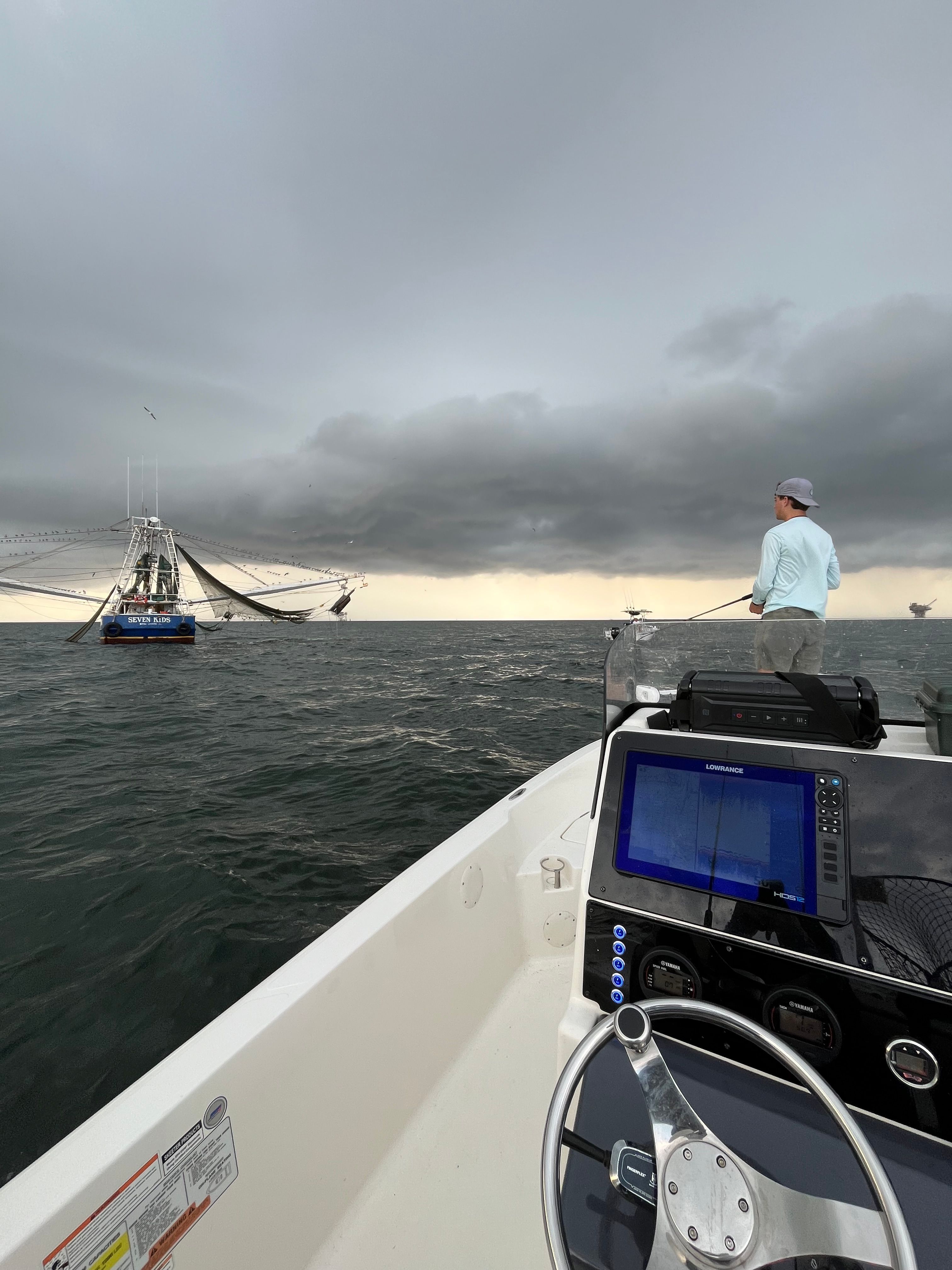 An angler stands on a boat on the water, with another boat nearby.