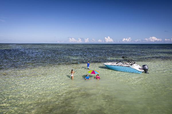2025 Hurricane SunDeck 235 OB boat anchored in shallow, clear water with people relaxing nearby.