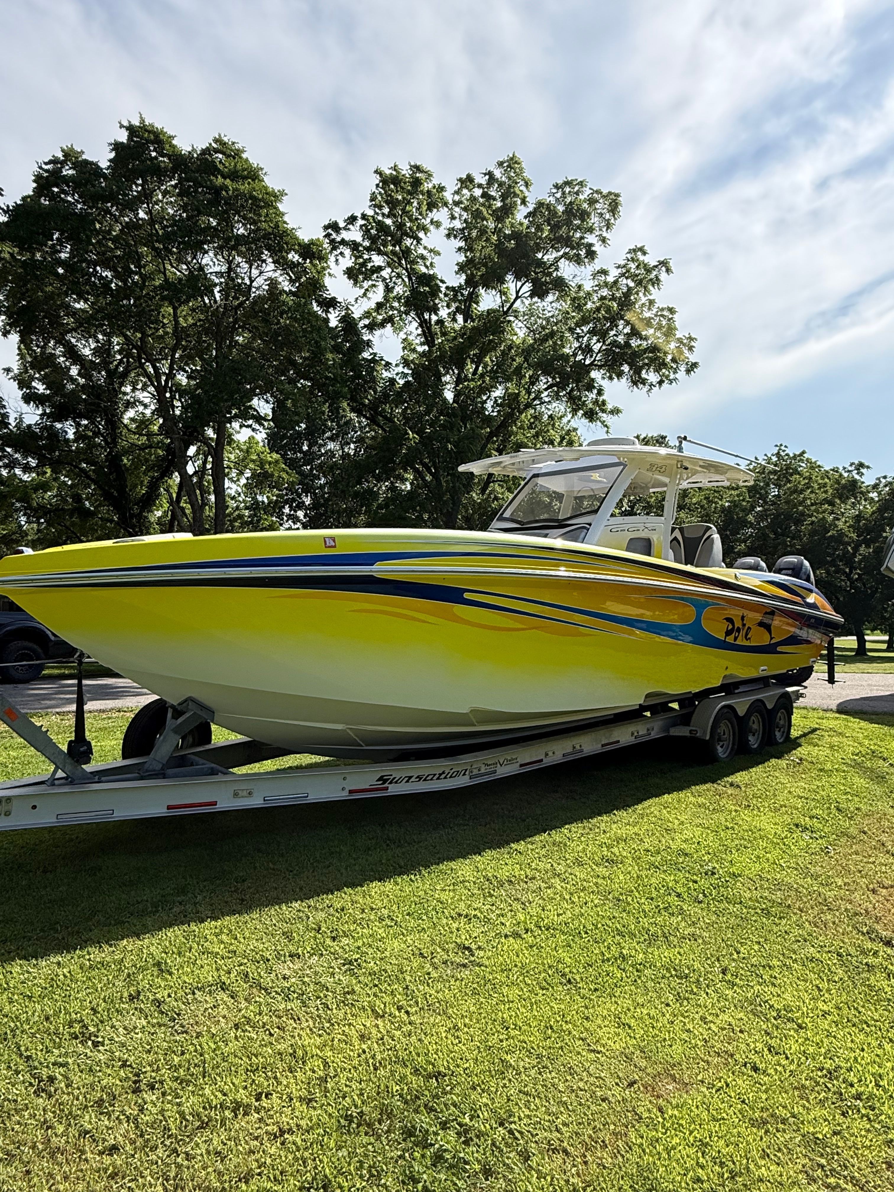 A yellow boat on a trailer on grass, surrounded by trees.