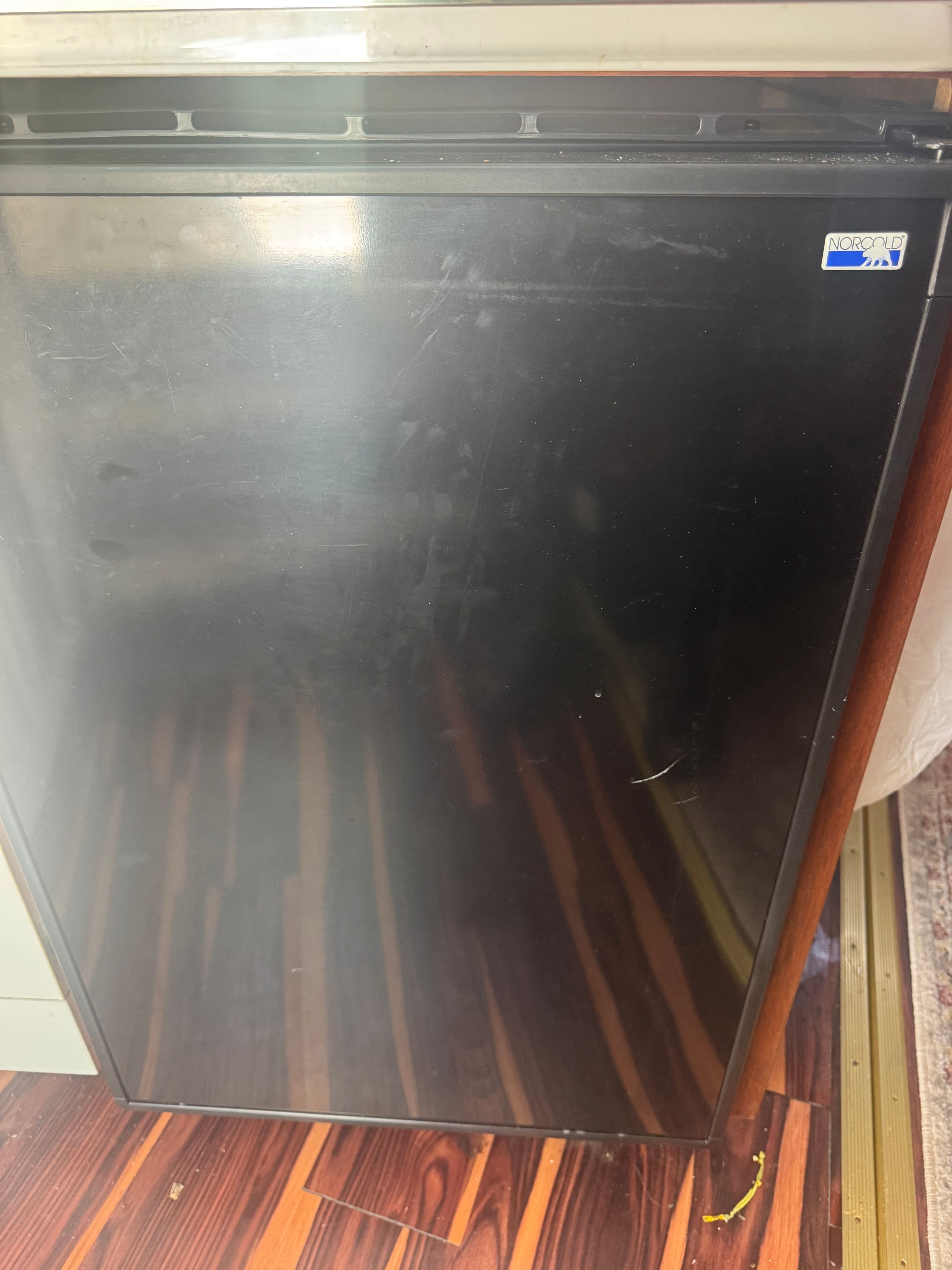 A compact black refrigerator stands on a glossy wooden floor inside a boat.