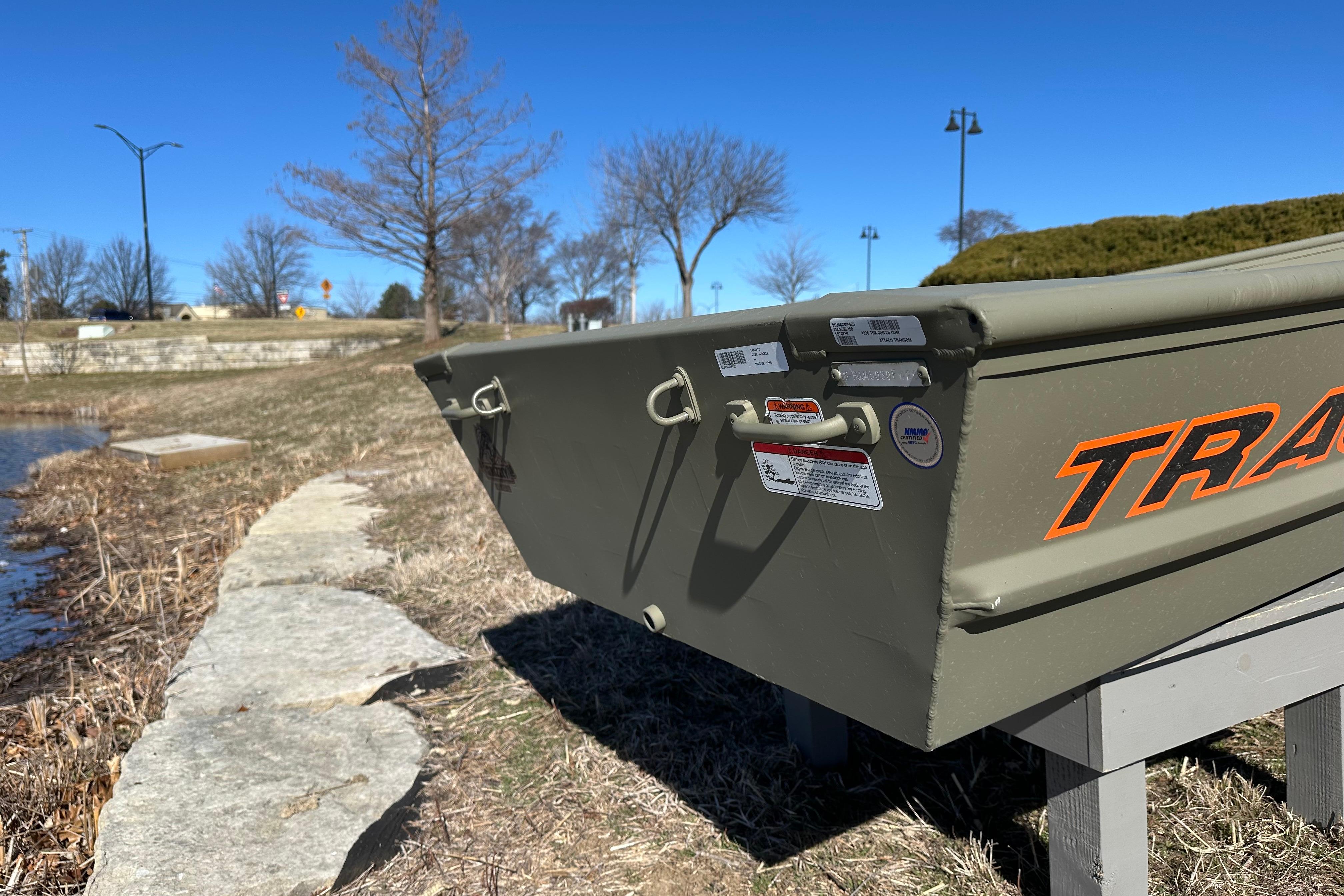 2026 Tracker Grizzly 12 Jon boat on grassy lakeside, clear blue sky background.