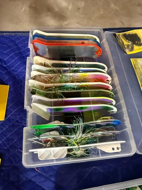 A box of various fishing lures, including colorful spoons and spinners, on a blue cloth.