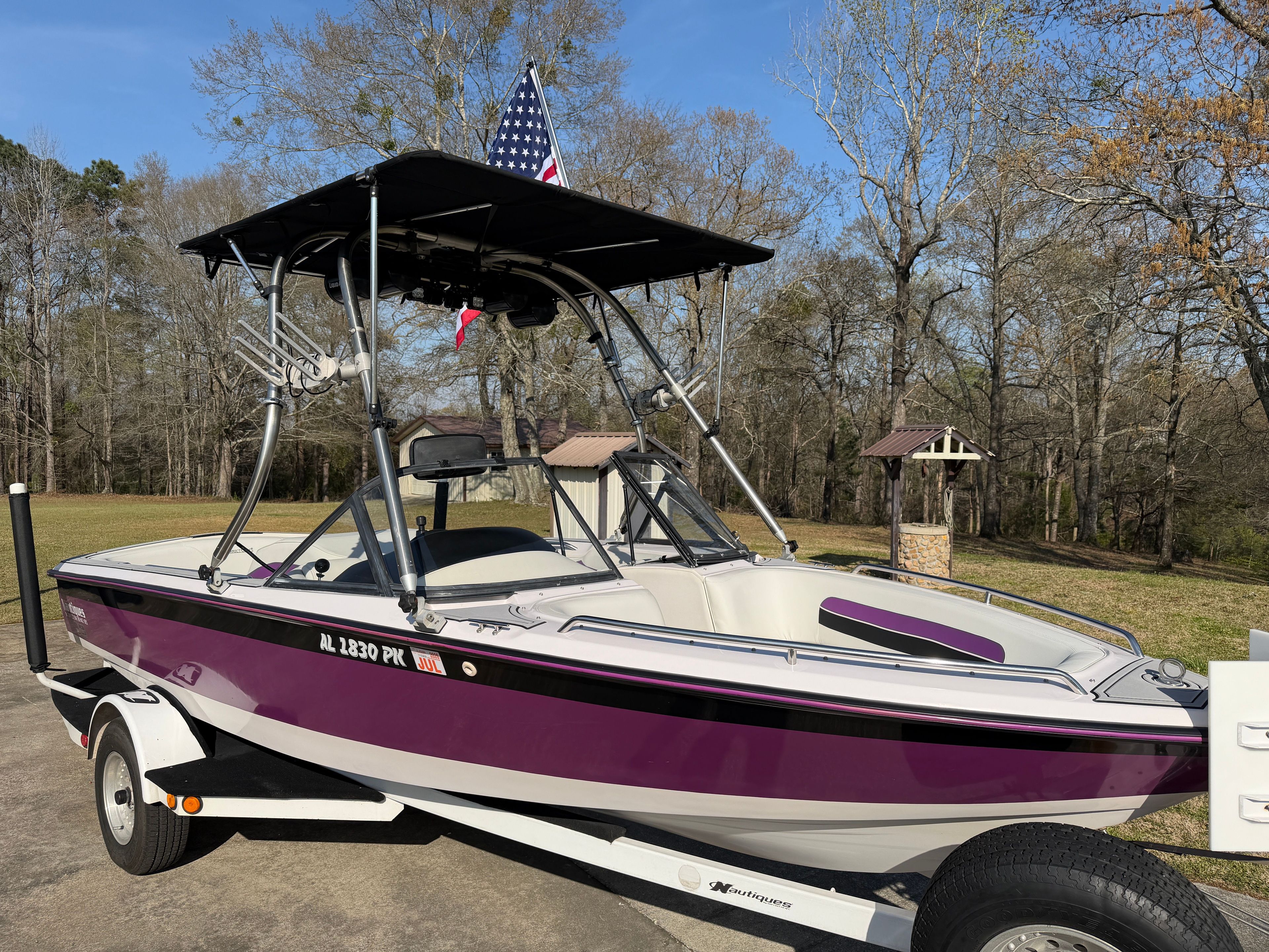 A purple and white sport boat with a black sunshade on a trailer in a wooded area.