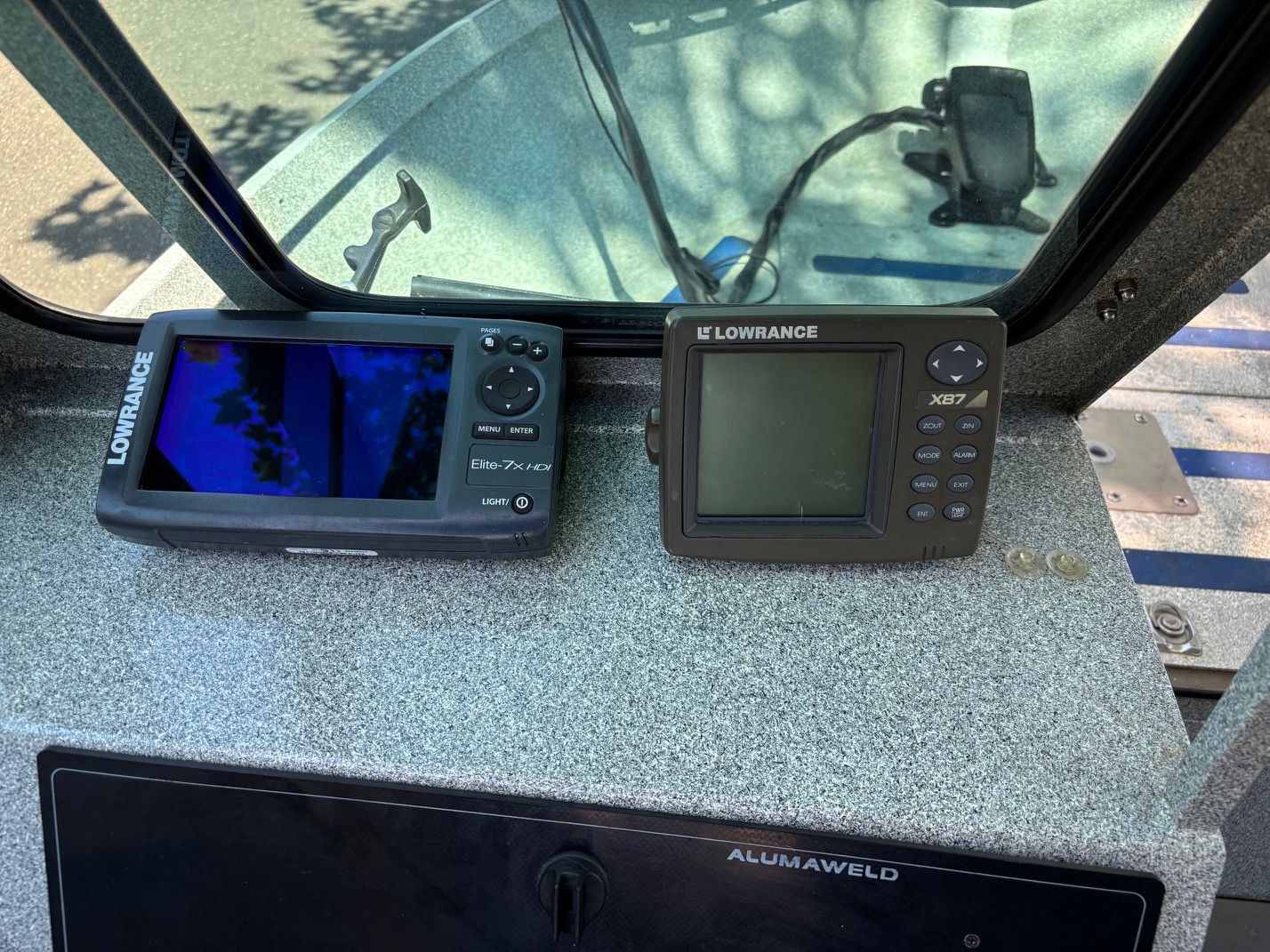Two Lowrance fish finders on a boat dashboard, surrounded by gray surface and a window.
