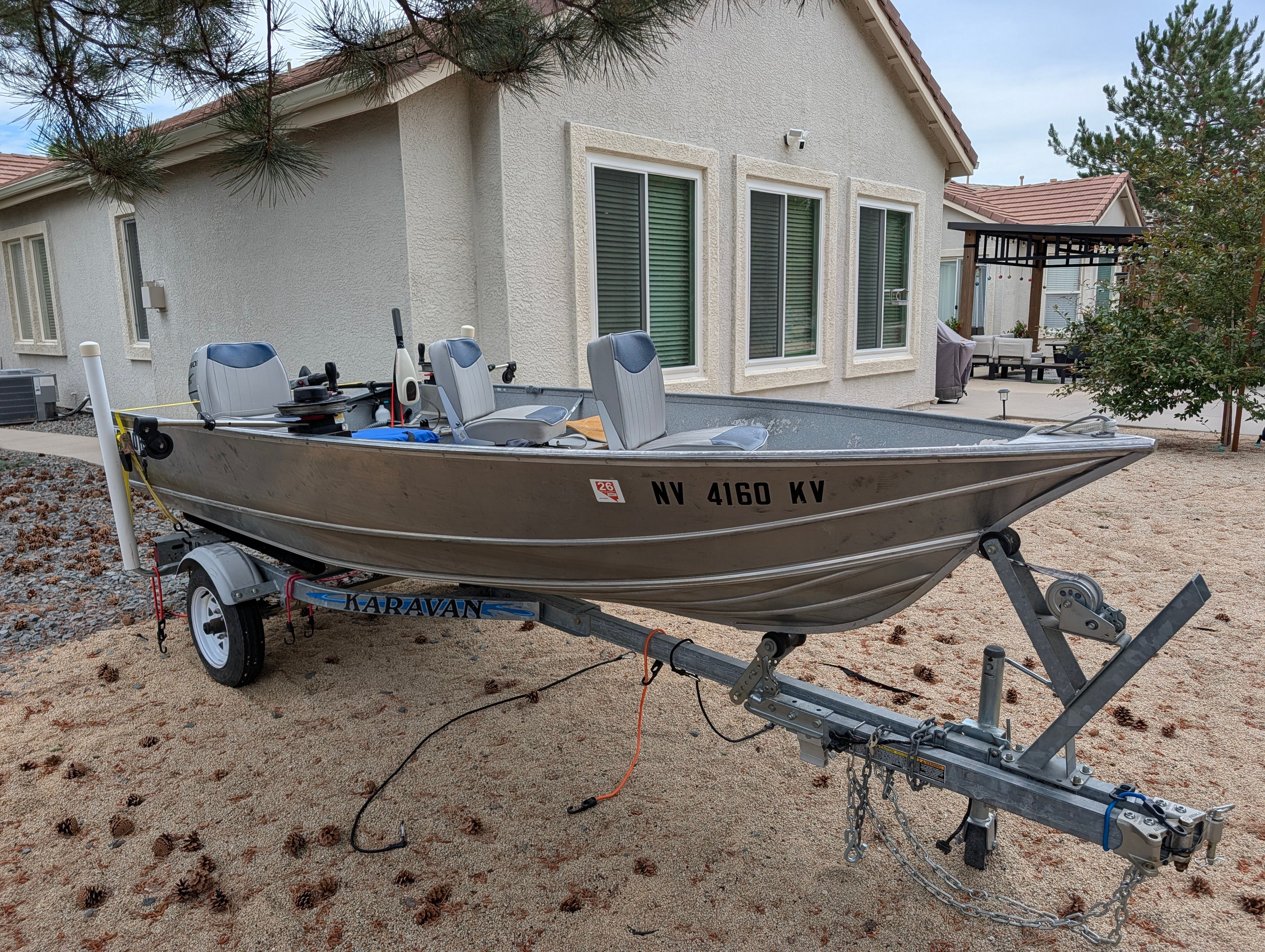 A silver aluminum boat on a trailer, parked on a sandy lot in front of a house.