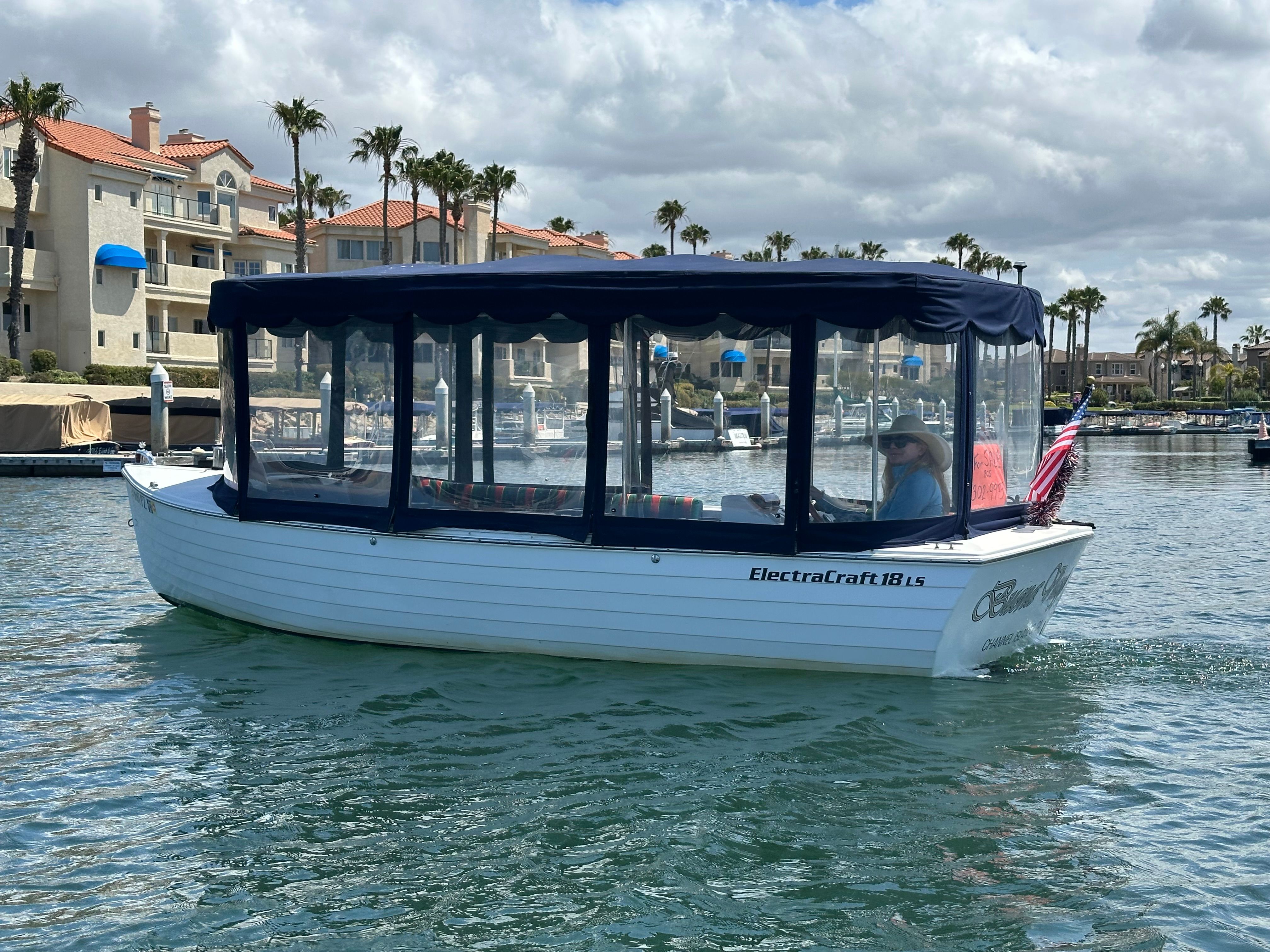 A white boat with a blue canopy cruises on calm water. Buildings and palm trees in the background.