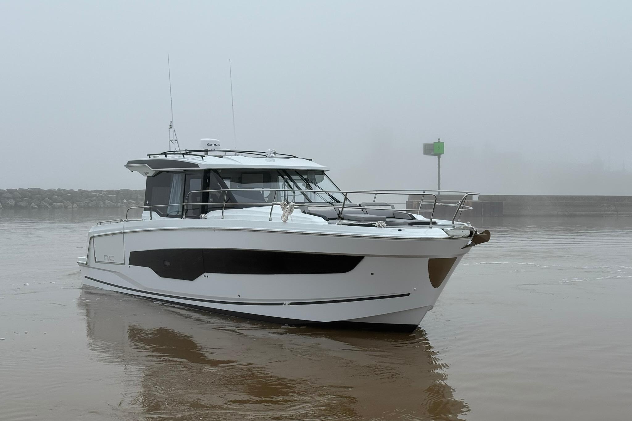 2026 Jeanneau NC 1295 Coupe boat on misty water near a rocky shoreline.