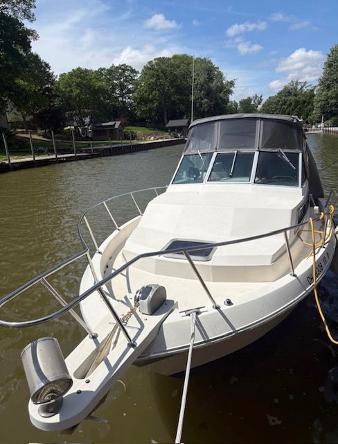 A white boat is docked at a calm riverside, surrounded by trees and houses.