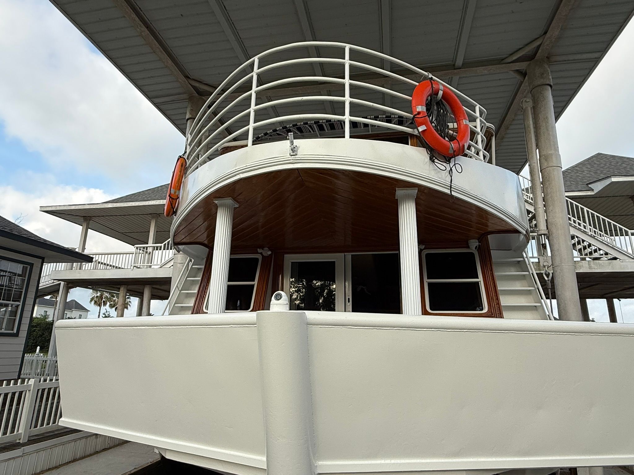 A two-story boat with white railings and lifebuoys, docked under a covered area.