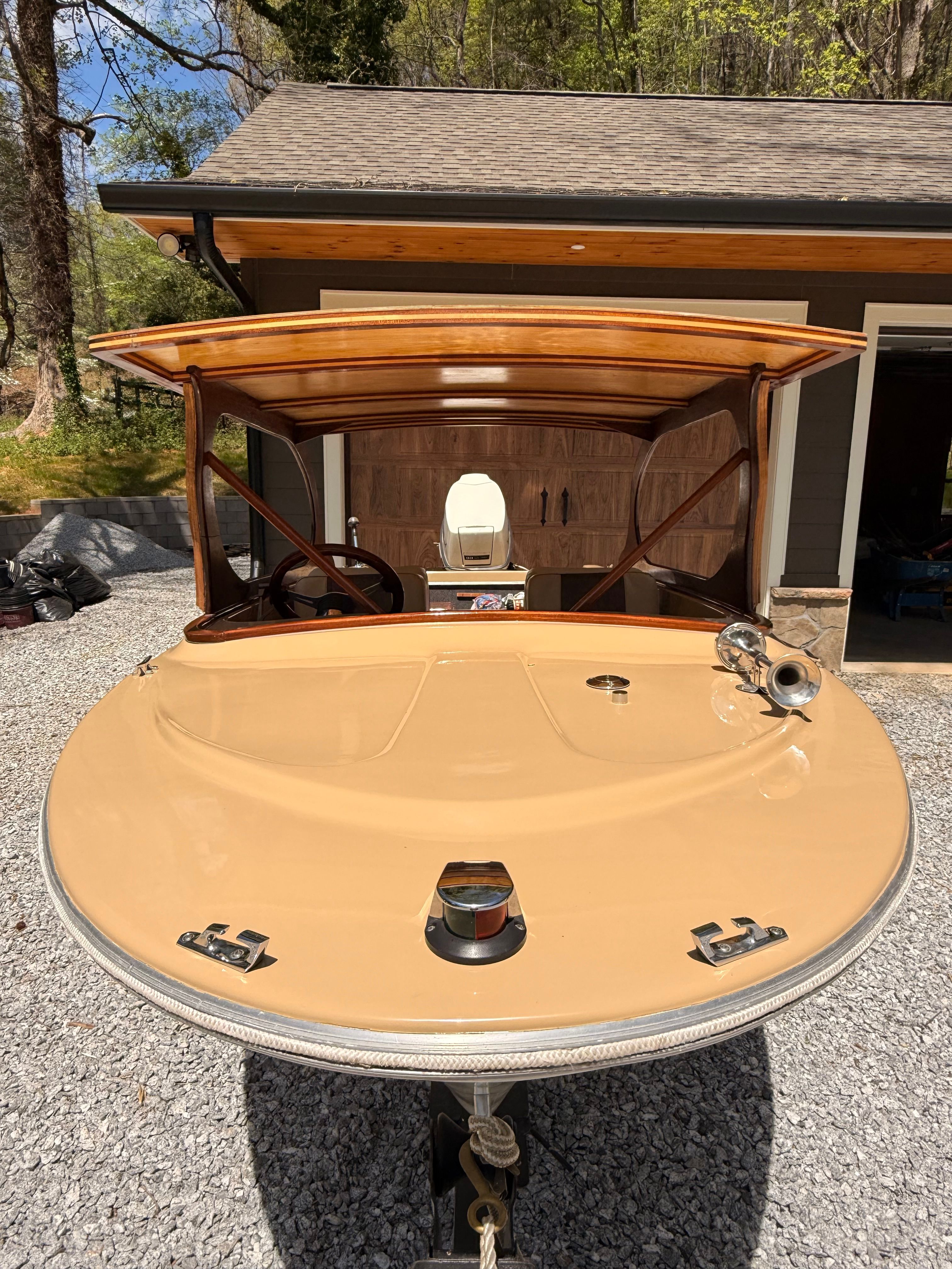 A vintage boat with wooden trim is parked on a gravel driveway in front of a garage.