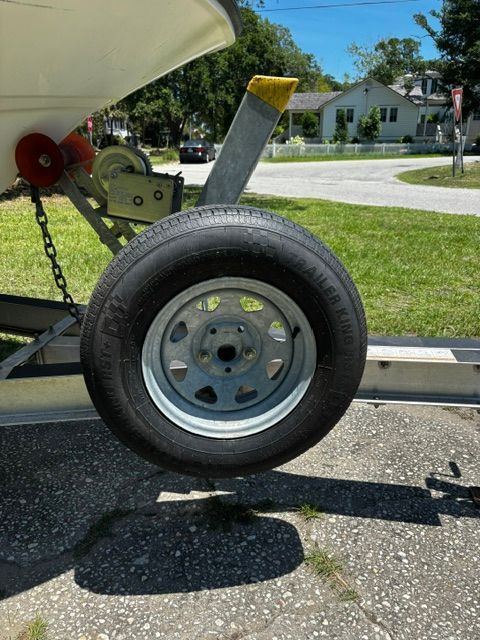 A boat trailer with a spare tire on a paved path, surrounded by grass and trees.