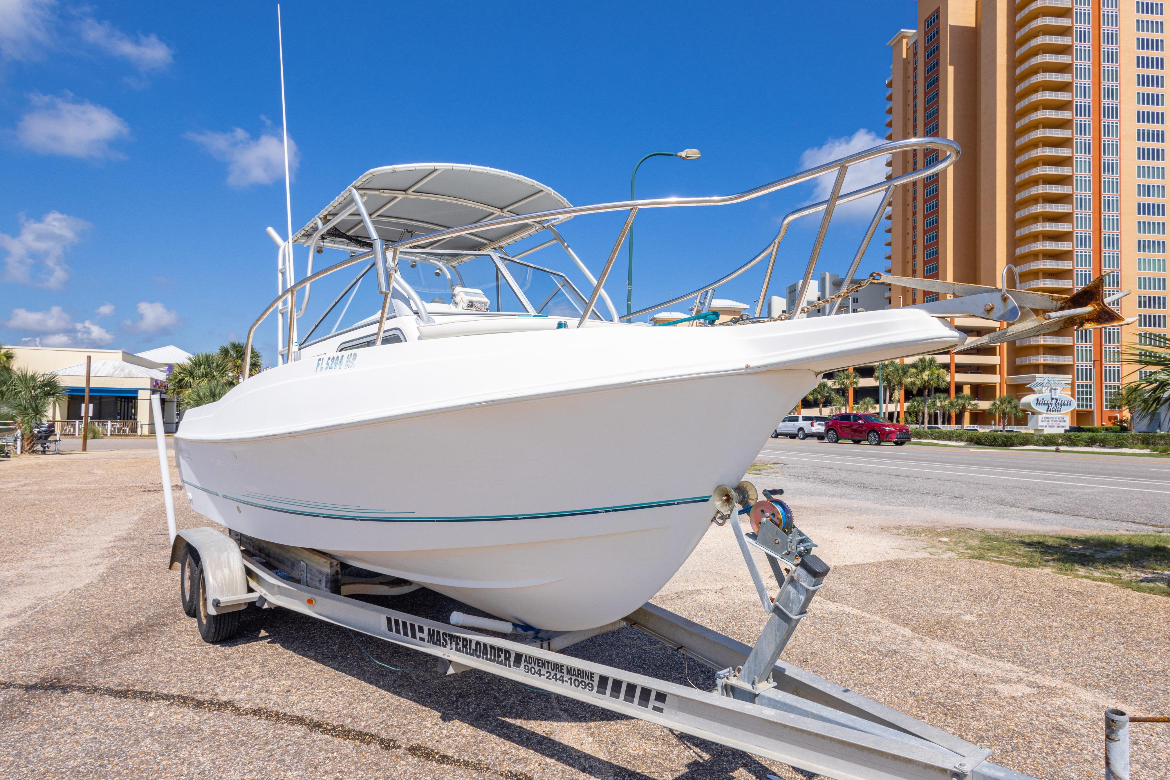 1999 Aquasport 225 Explorer boat on trailer, parked near tall buildings under a clear blue sky.